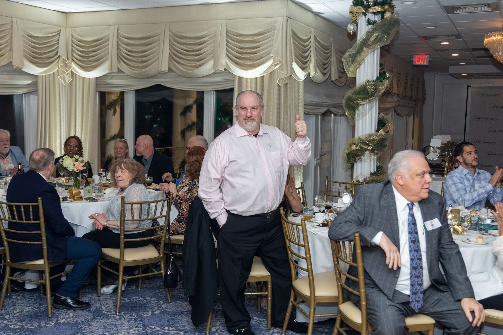 Man giving thumbs-up at a formal event, standing near a table with people.