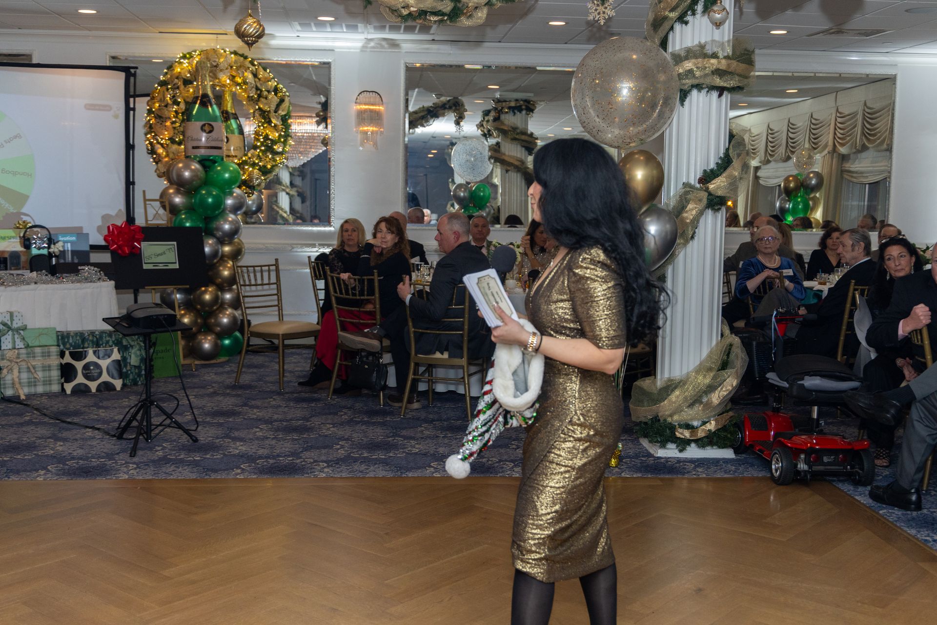 Woman in gold dress at a decorated event, holding a gift, speaking to guests seated at tables.