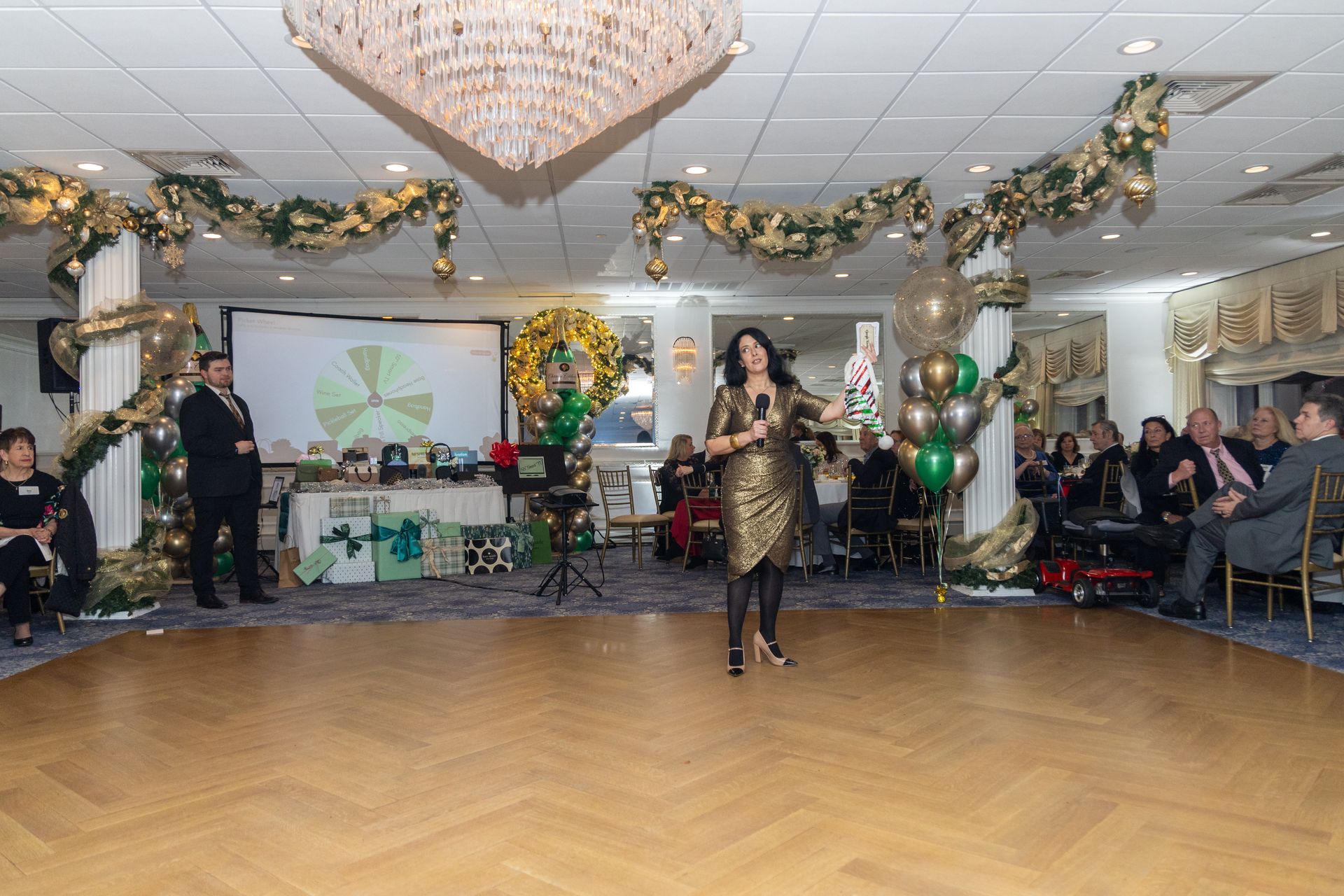 Woman in gold dress speaks at event with green and gold decorations.