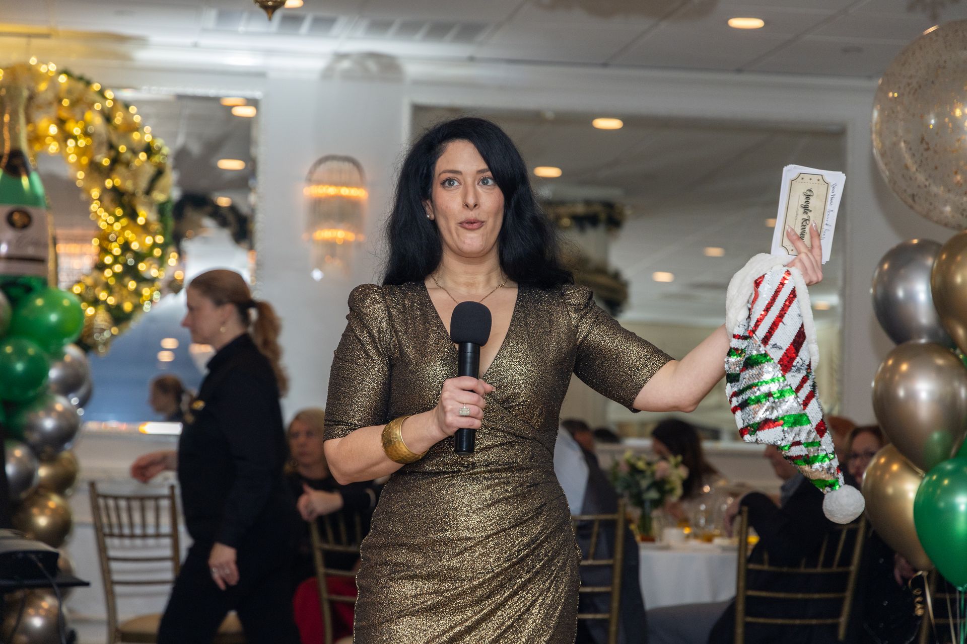 Woman in gold dress holding a Christmas stocking, speaking into a microphone at an event.