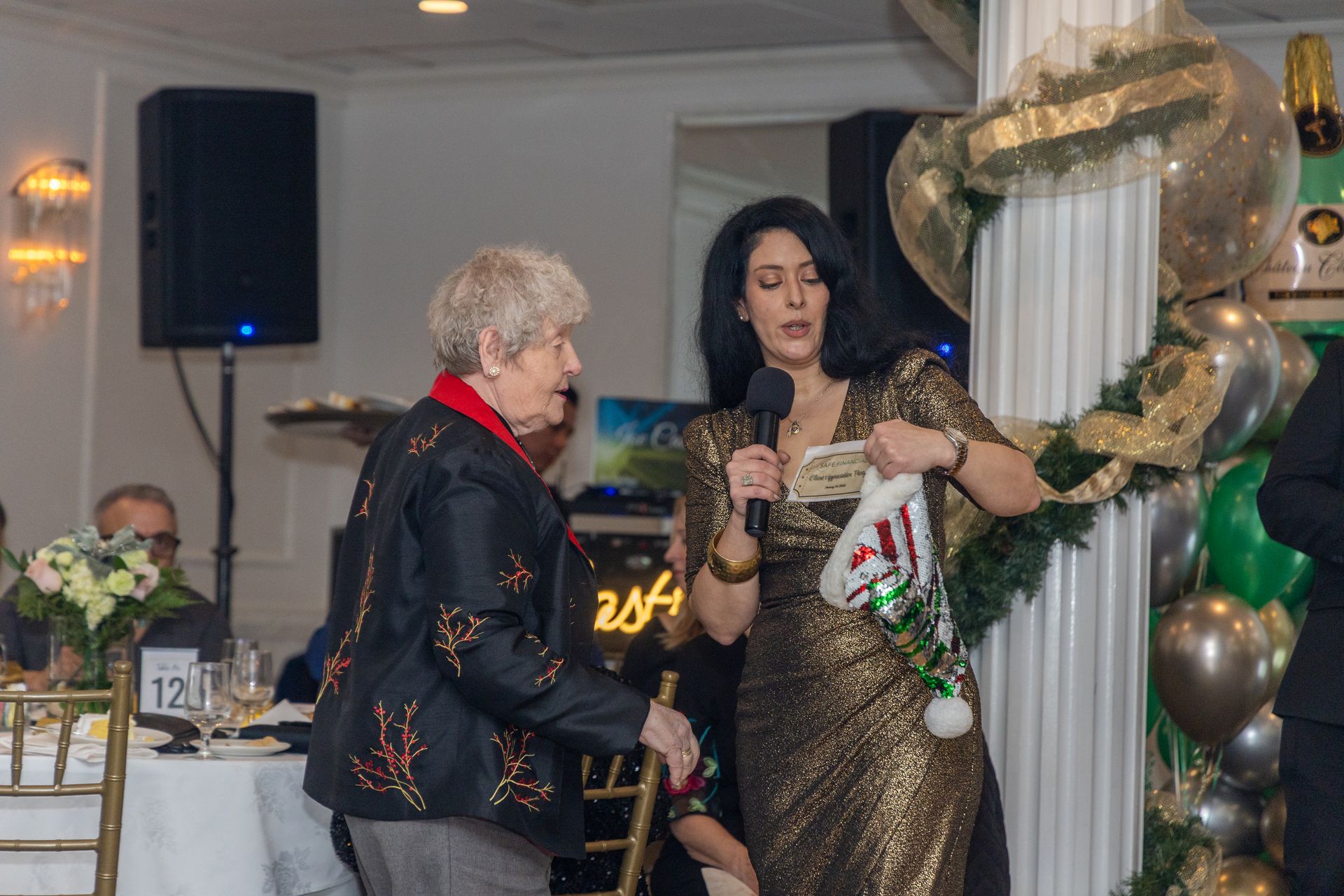 Woman in gold dress holds a Christmas stocking, speaking to an older woman at a party.