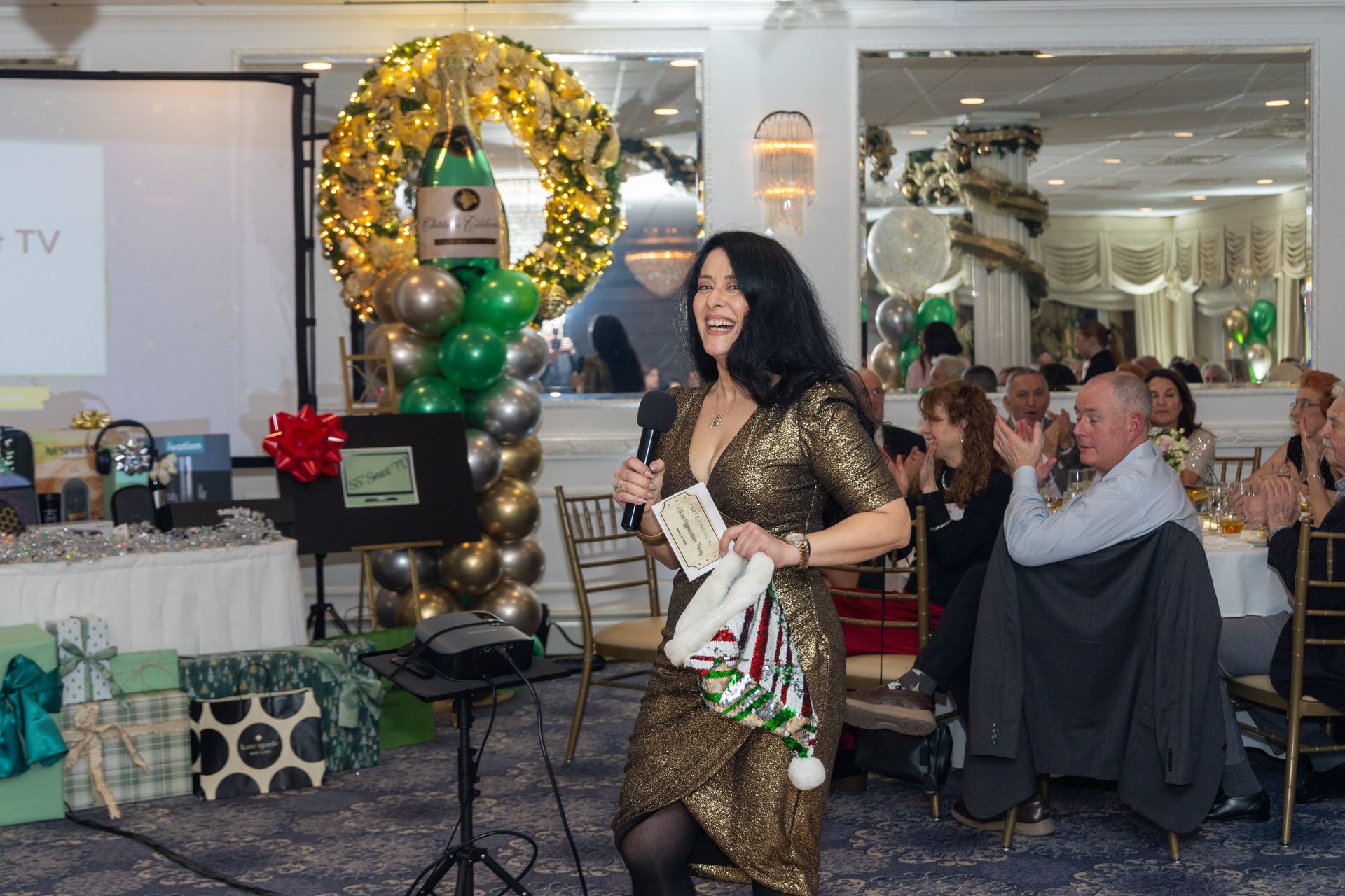 Woman in gold dress speaks at event, holding microphone and Christmas hat, festive decorations behind.