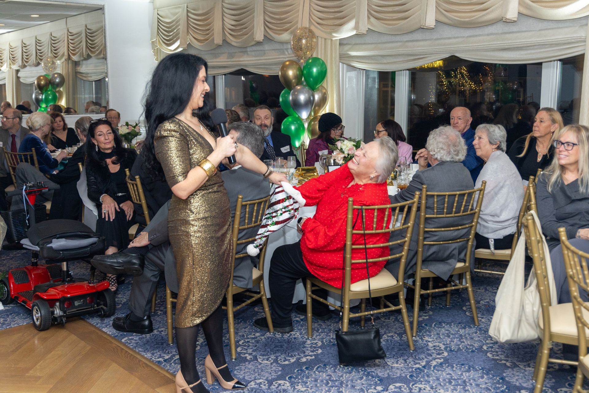 Woman in gold dress speaks into microphone at event; seated guests listen.