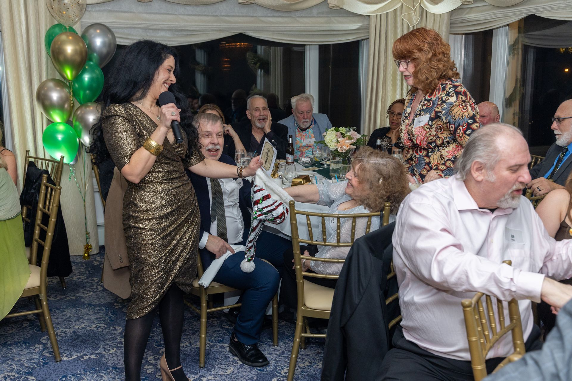 Woman in gold dress speaks into mic at a party. People seated at tables laugh. Balloons.