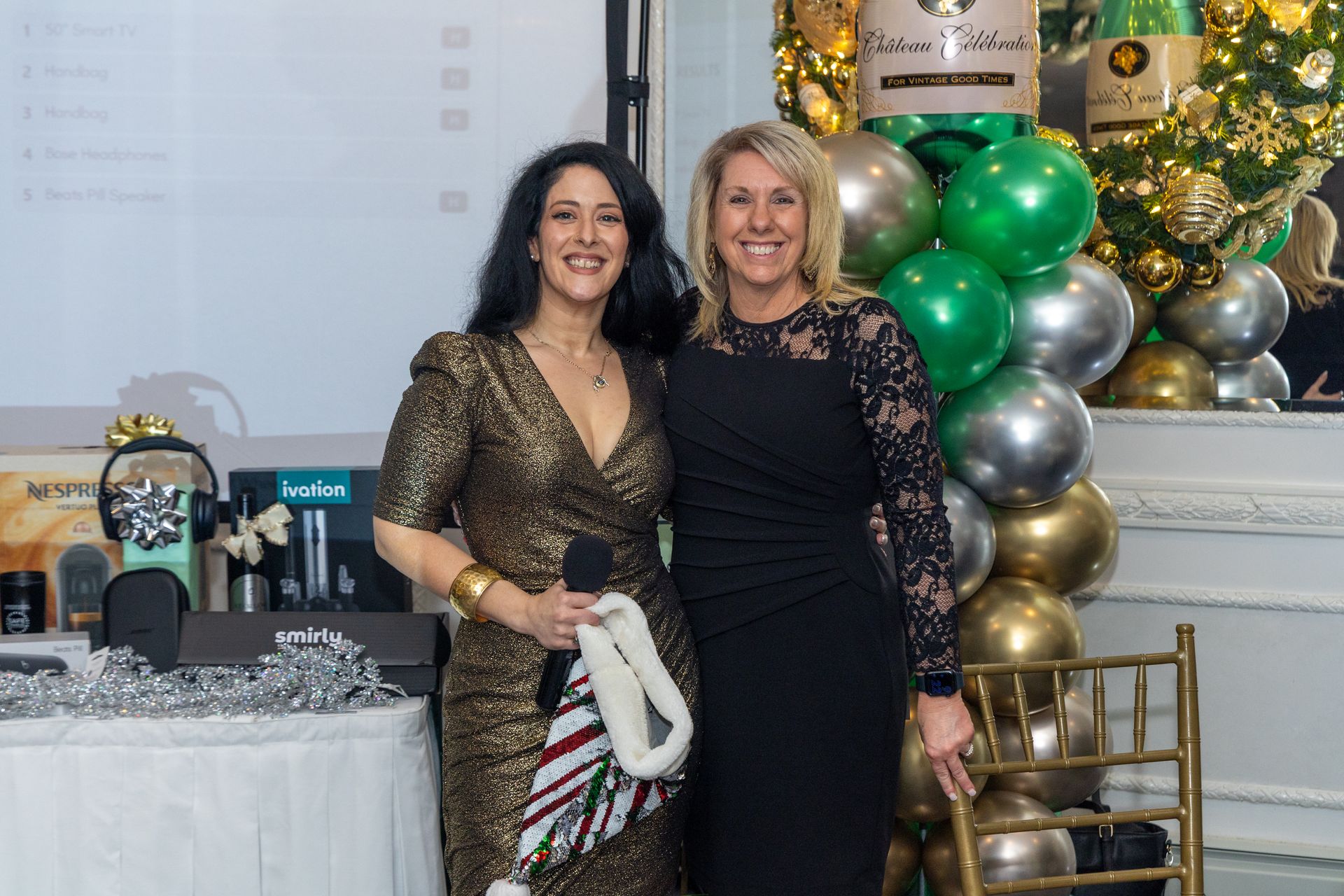 Two women smiling, posing at event. Woman in gold dress holds microphone, other in black dress. Festive balloons.