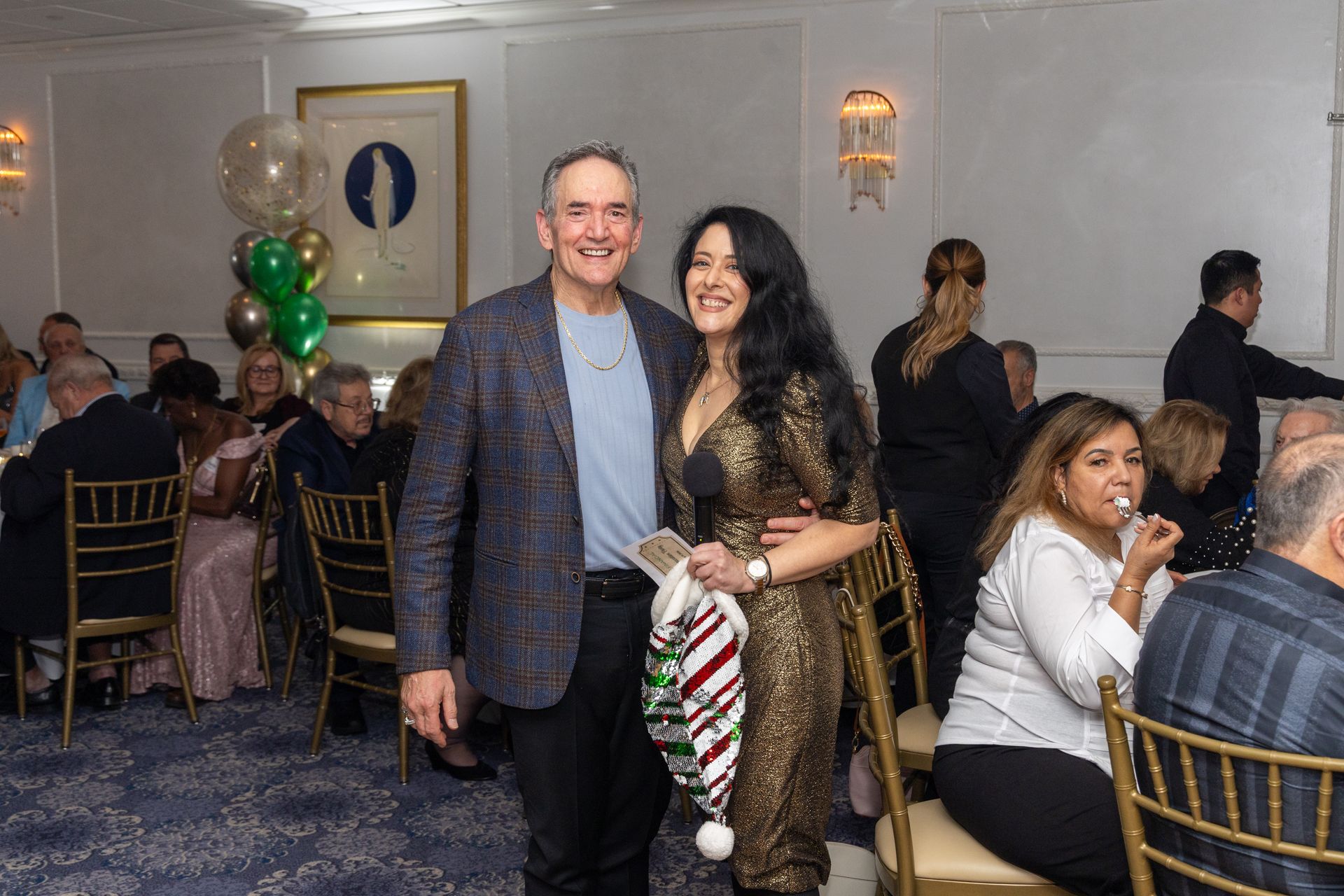 Couple smiles, poses at an event, man in blazer, woman in gold dress, seated guests in background.