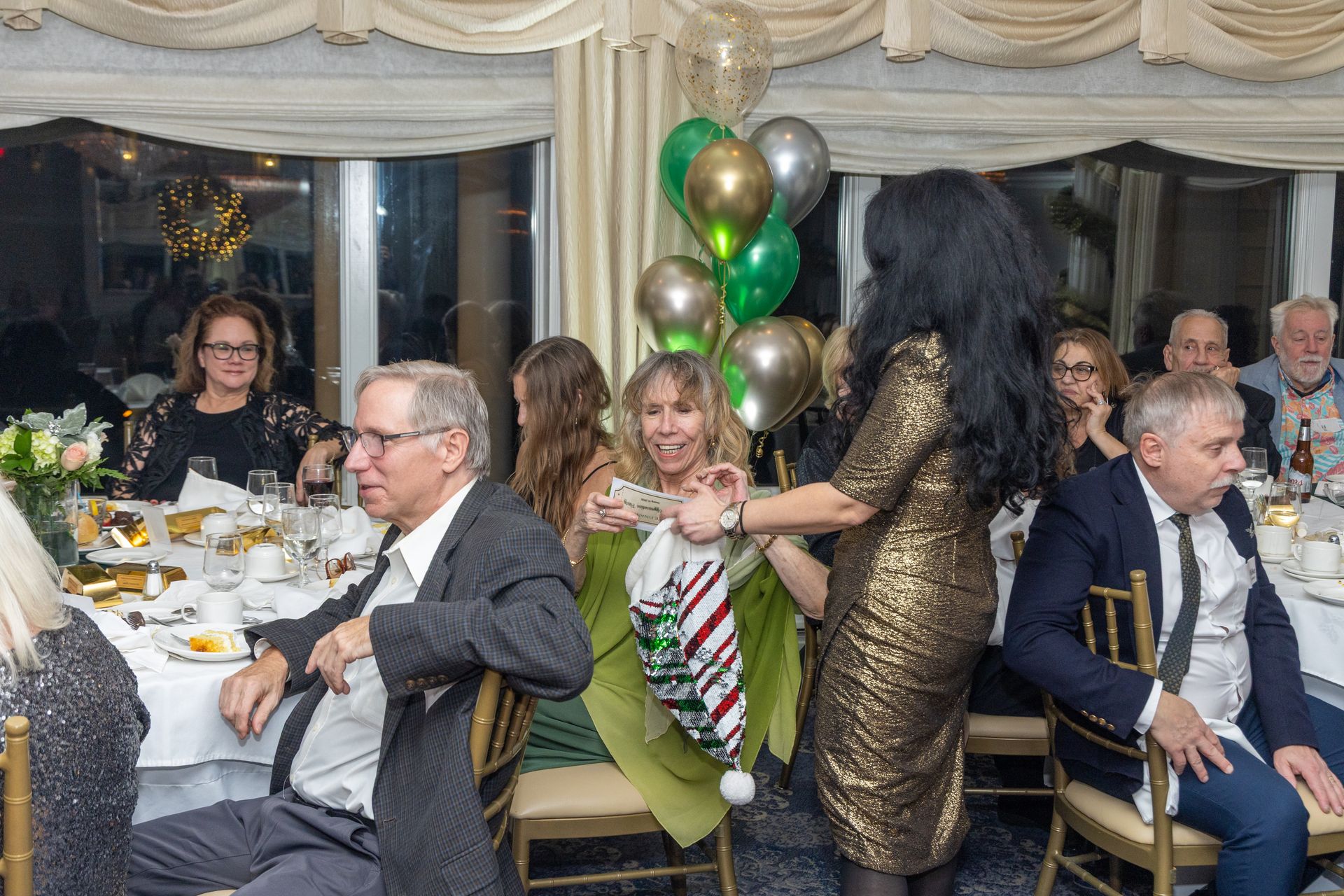 People at a formal event, some seated at tables with food, others interacting. Festive decorations are present.