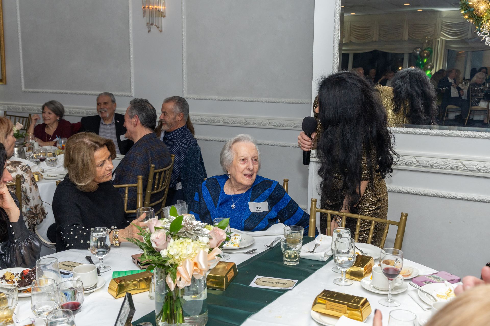 Woman speaking into microphone at a table during an event. Guests at tables, gold bars on the table, floral centerpieces.