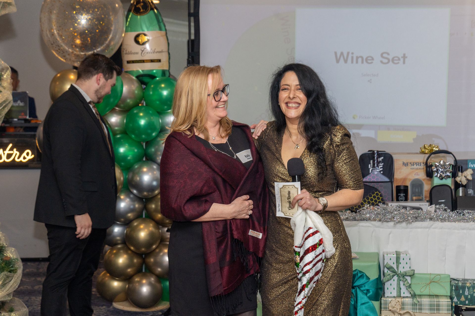 Two women smiling on a stage, one holding a microphone and a paper. A wine set is on display.