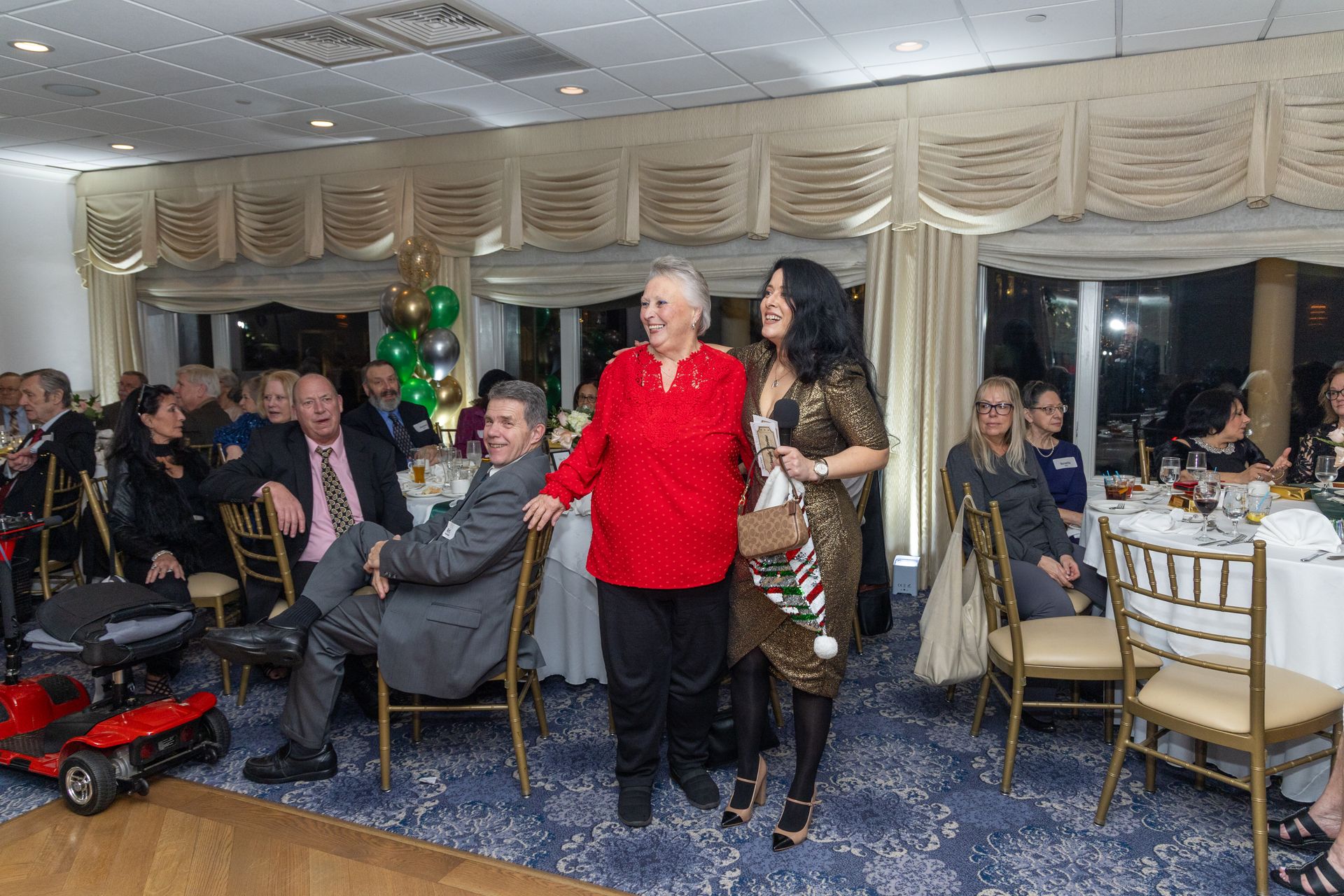 People at a festive event in a banquet hall. A woman in a red shirt stands with two people, smiling.