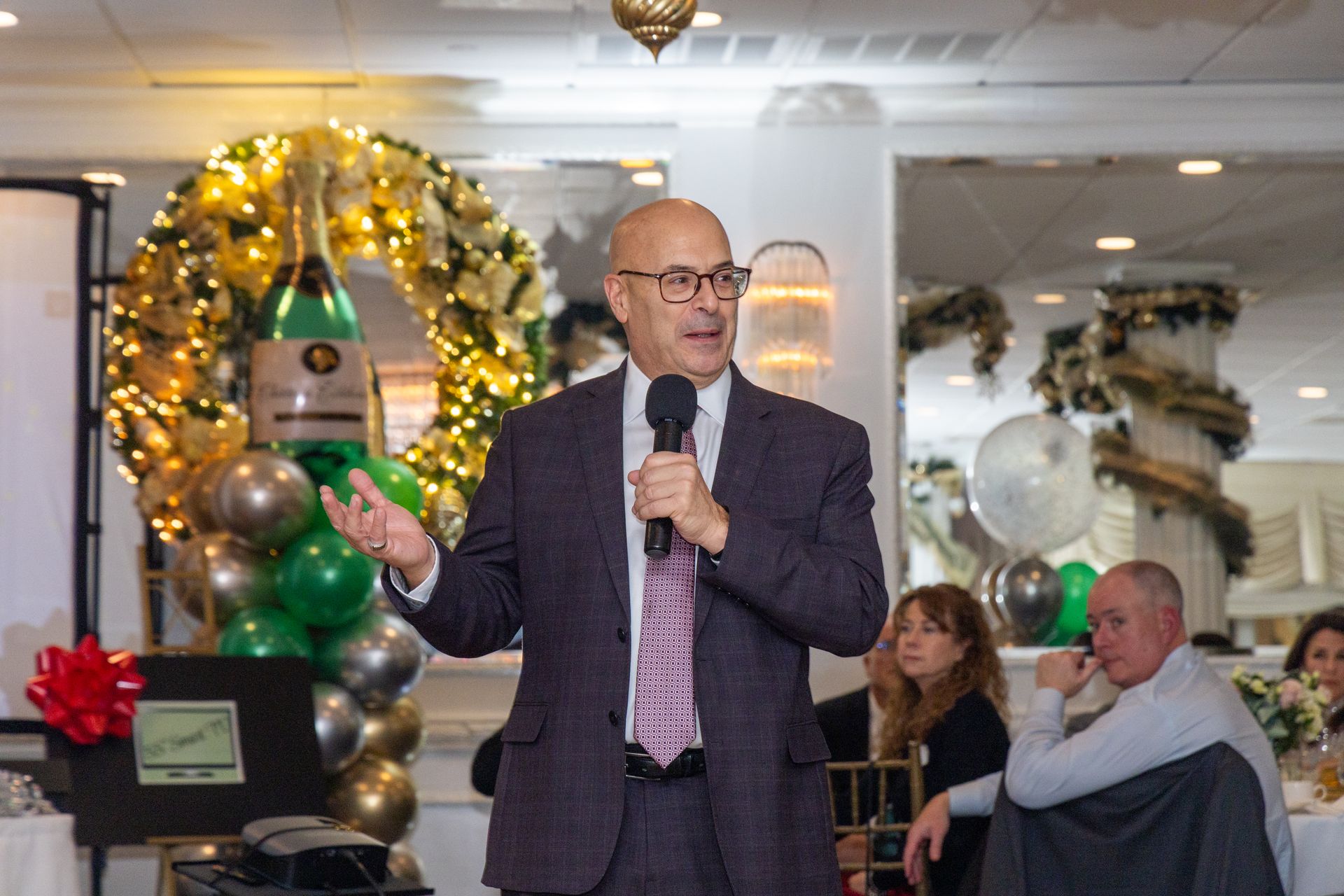 Man in suit speaks into microphone at event with decorations.