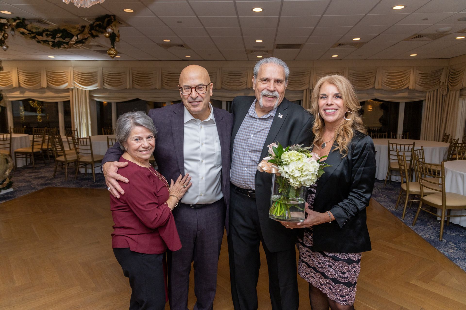 Four people pose together indoors, smiling, one holding flowers. Formal attire.