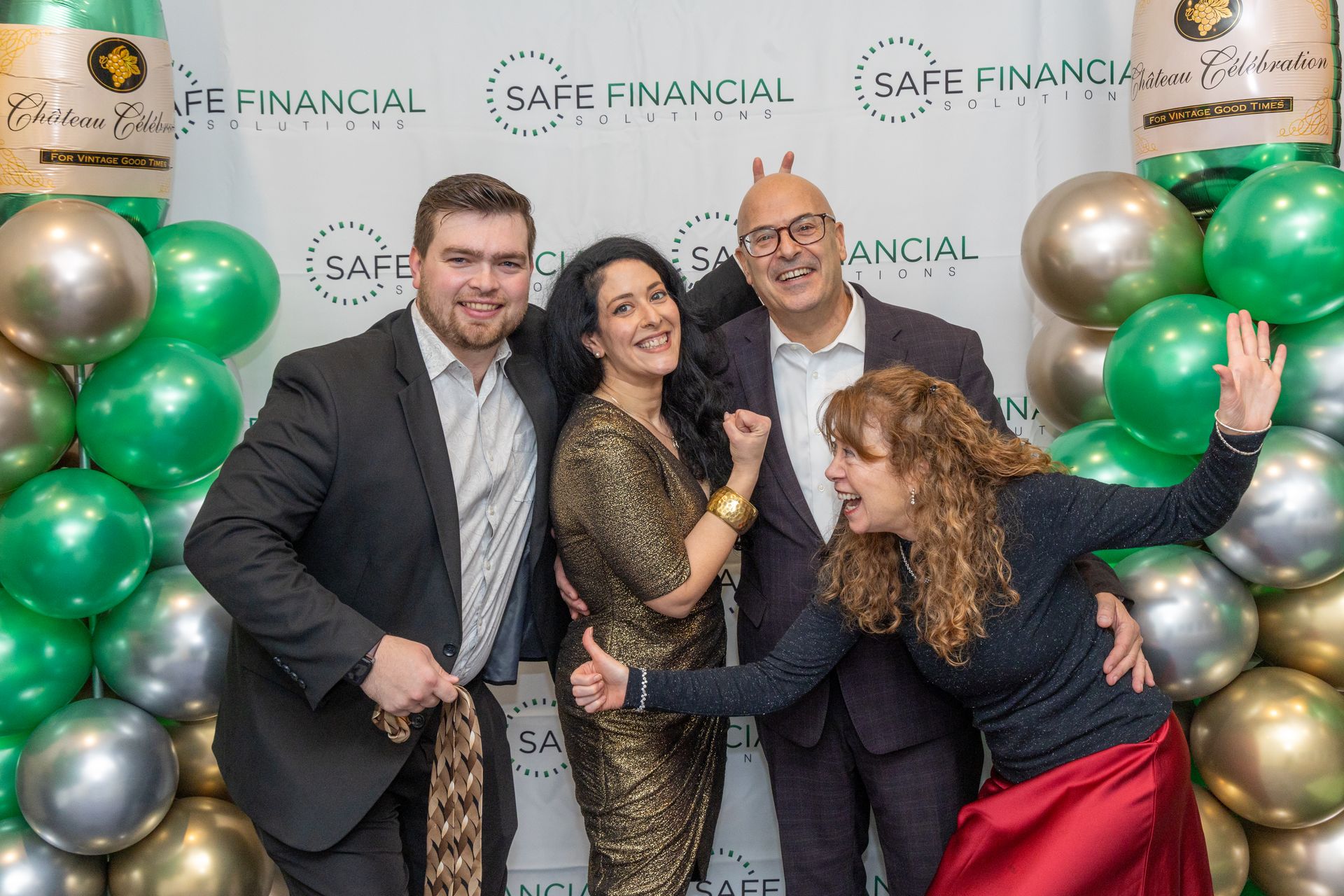 Four people pose playfully in front of a backdrop with balloons. One makes bunny ears, others smile.