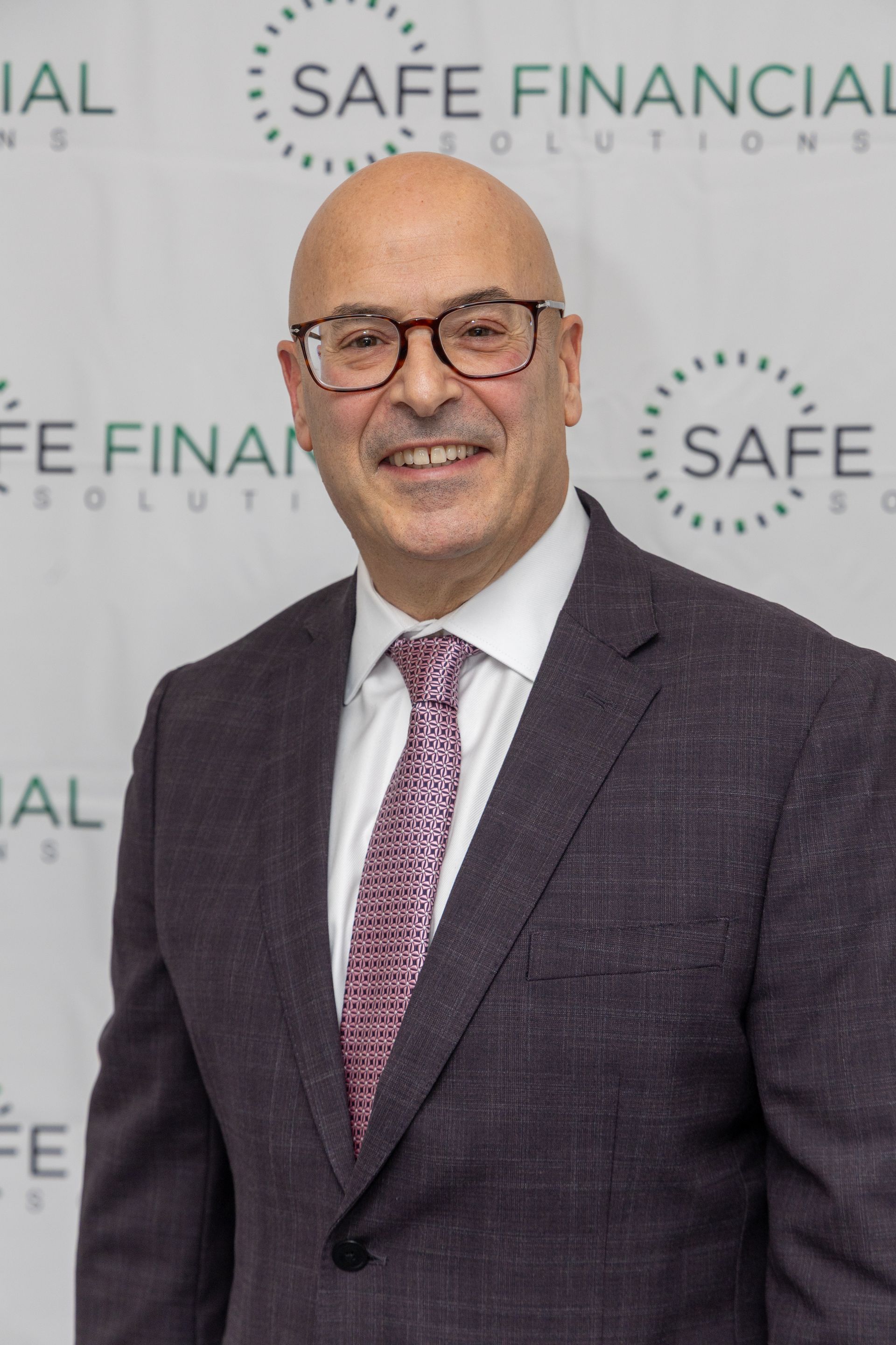 Bald man in glasses wearing a suit and tie, smiling in front of a company logo backdrop.