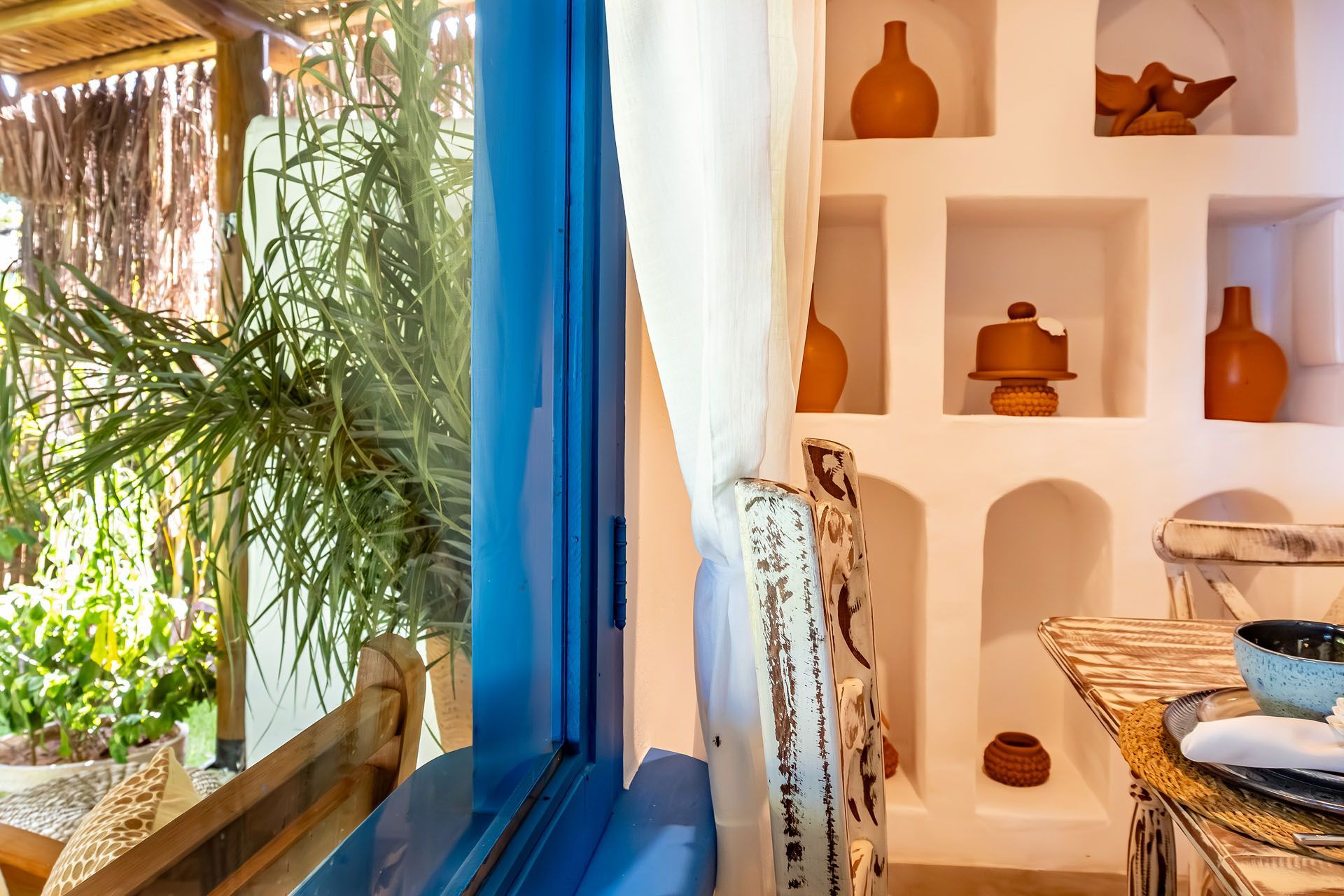 Quarto boêmio e luminoso com cortinas azuis, prateleiras brancas com cerâmica e uma planta junto à janela aberta.