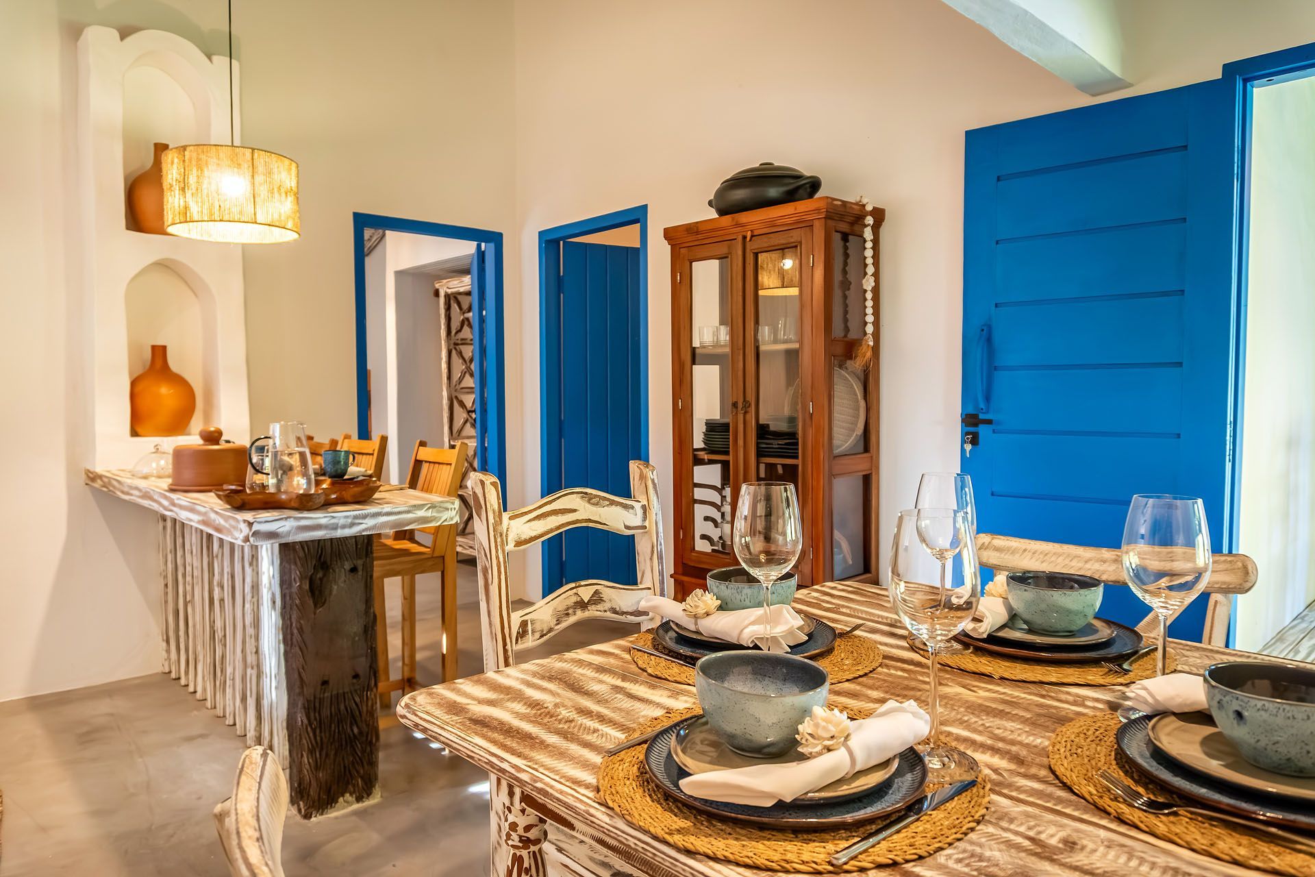 Sala de jantar iluminada com portas azuis, mesa de madeira posta para refeições e um aparador com louças.
