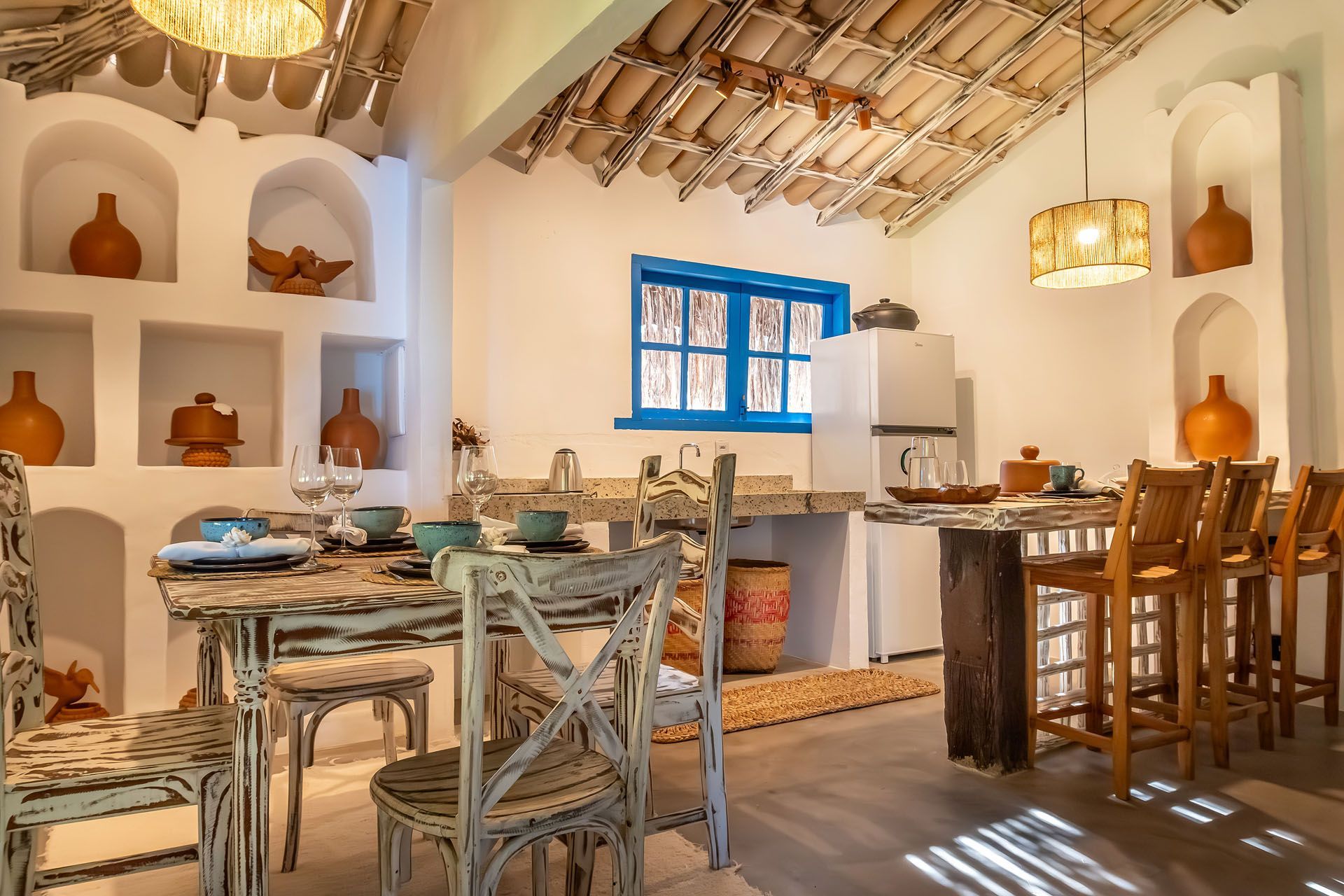 Sala de jantar rústica com mesas de madeira, molduras azuis nas janelas, cadeiras de vime e prateleiras embutidas brancas com peças de cerâmica.