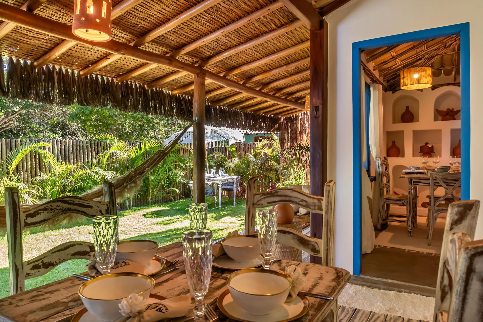 Área de jantar rústica ao ar livre com mesa posta para dois, cadeiras de madeira e porta com moldura azul que dá acesso a um interior aconchegante.