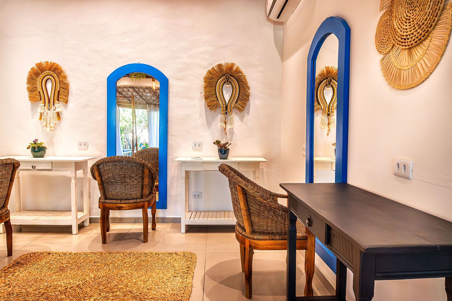 Quarto iluminado com espelhos com moldura azul, cadeiras de vime, escrivaninha branca, mesa preta e decoração de parede com palha.