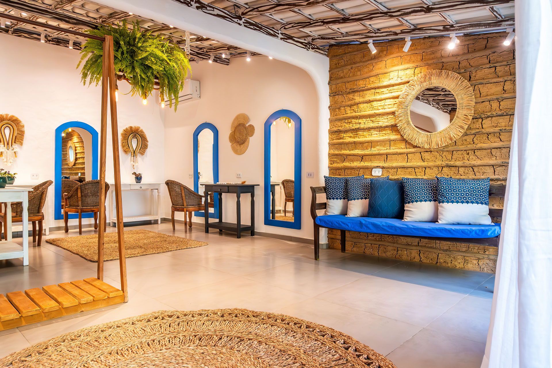 Quarto em estilo boho com detalhes em azul, plantas suspensas, decoração em tecido e um banco almofadado junto a uma parede de tijolos.