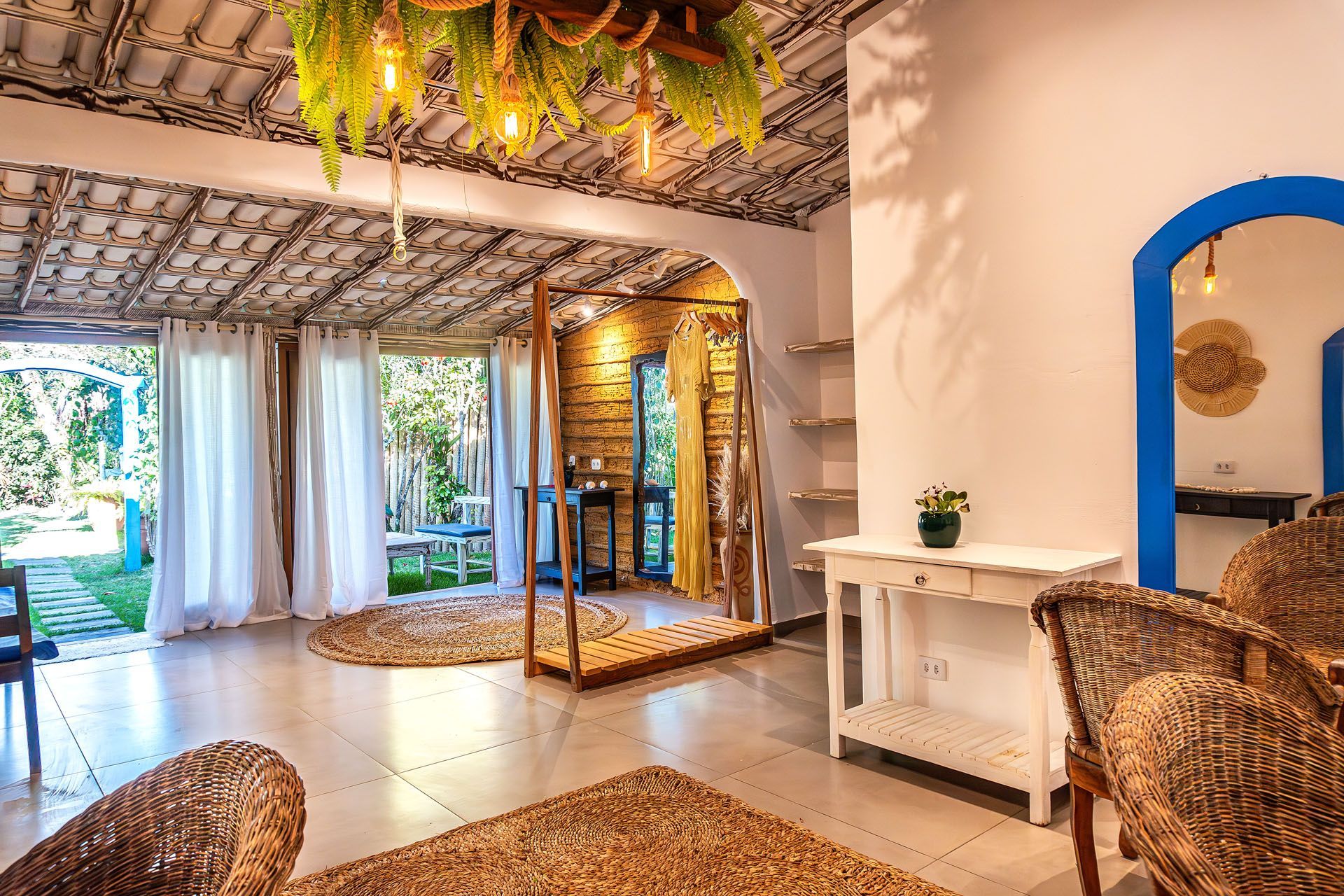 Quarto tropical luminoso com cadeiras de vime, espelho azul, plantas e portas abertas para um jardim ensolarado.