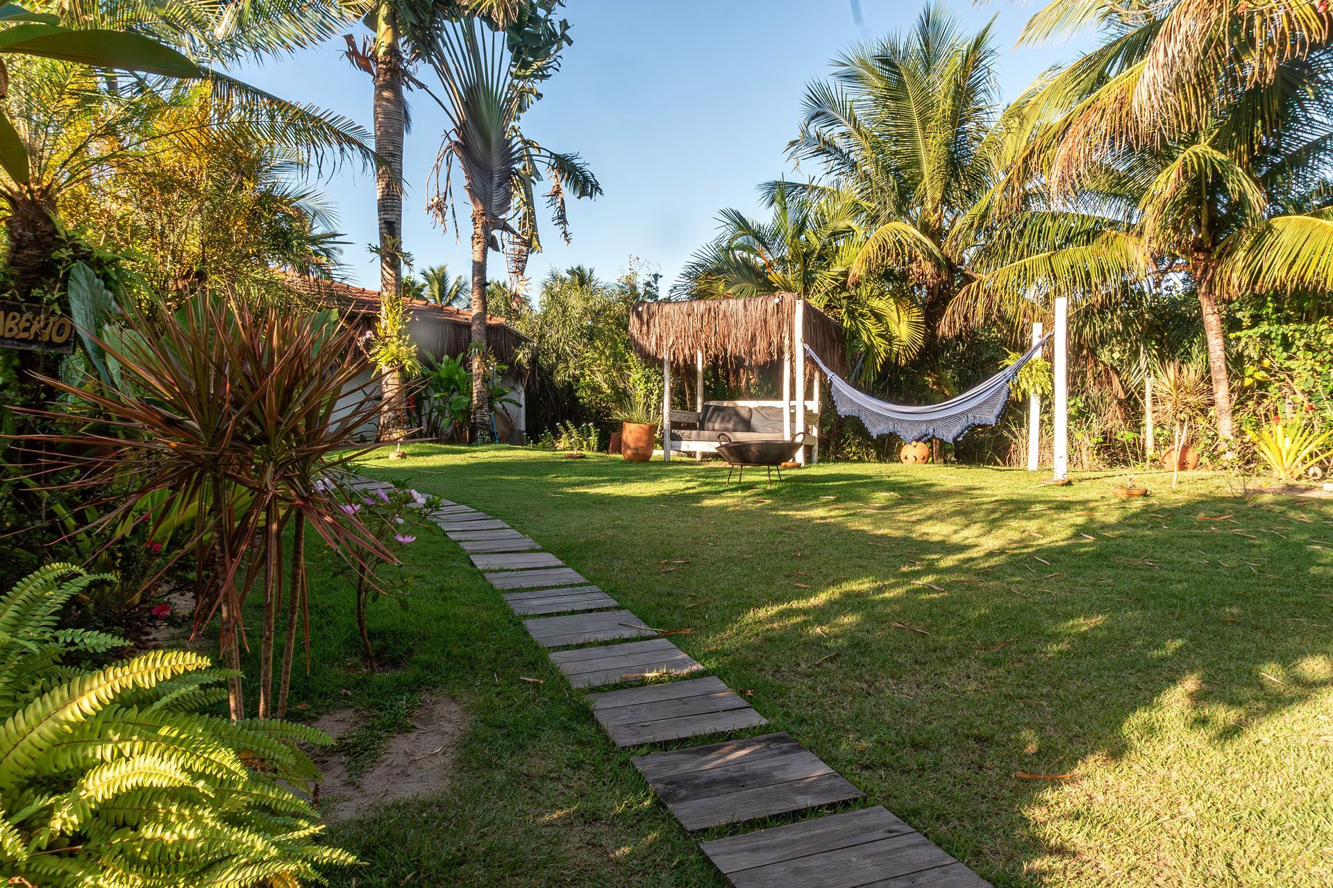 Jardim tropical ensolarado com caminho de pedra, gramado, palmeiras e uma rede junto a uma pérgola sombreada.
