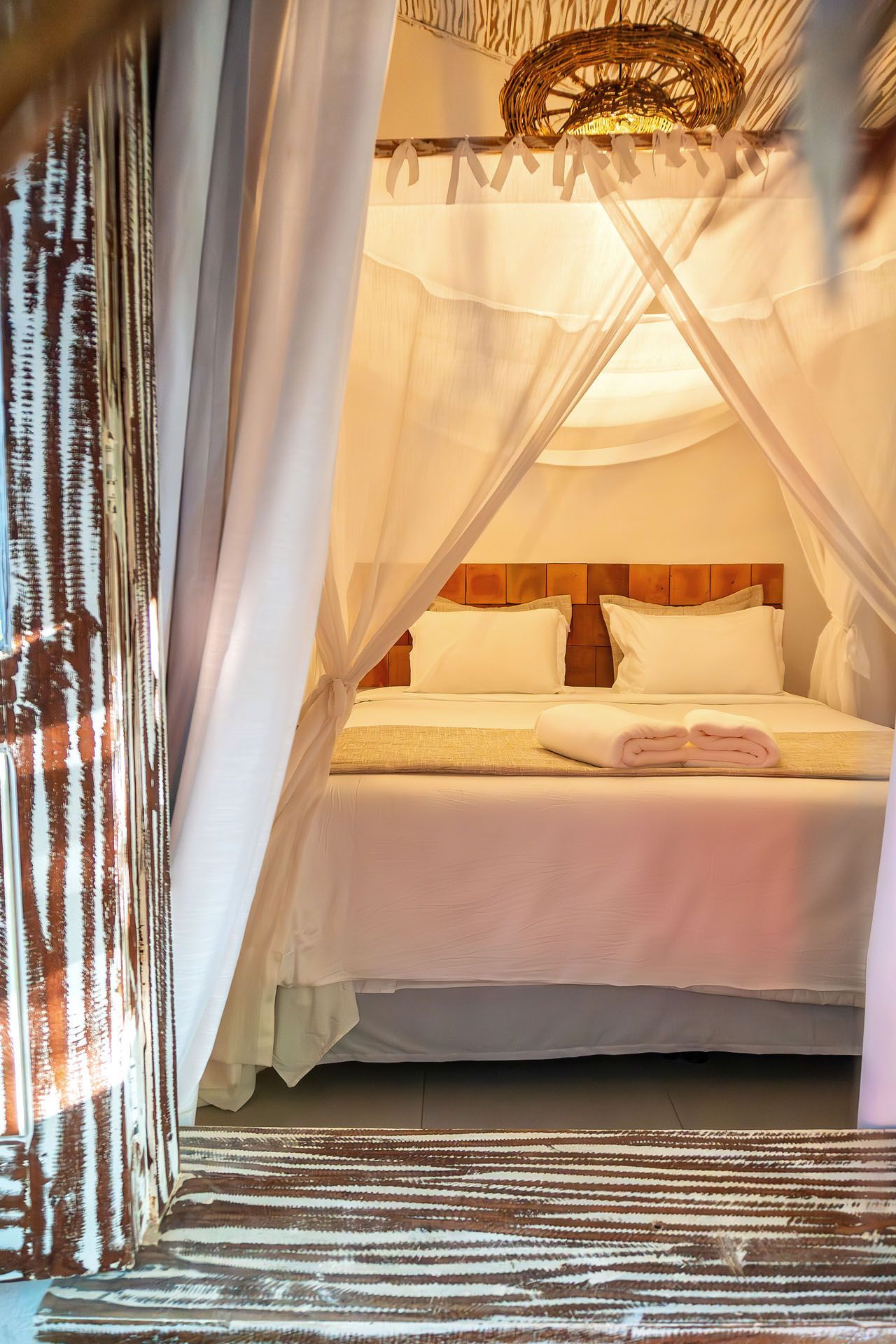 Quarto com cortinas leves, cama branca, iluminação aconchegante e tapete estampado no chão.