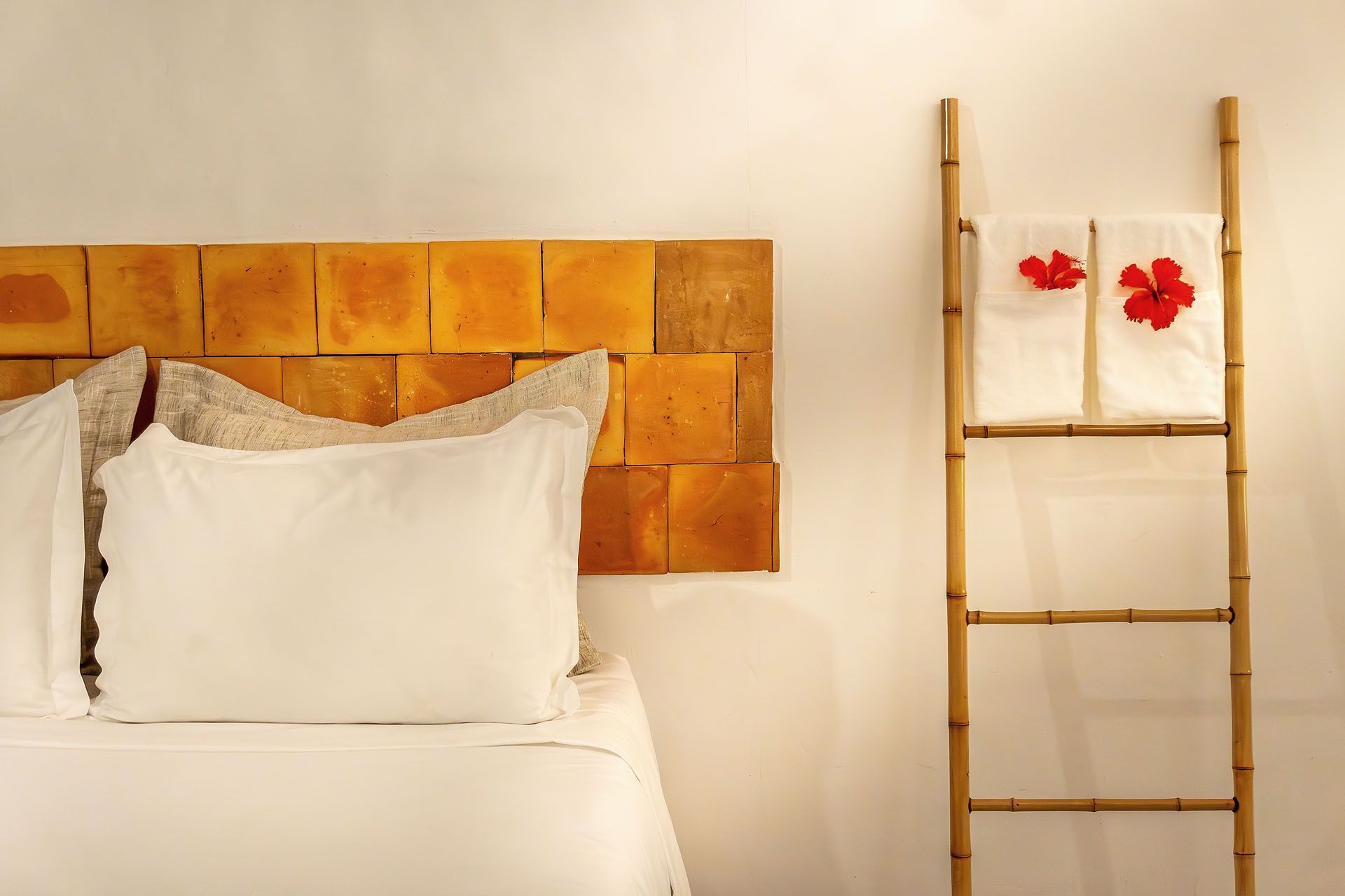 Cama branca com cabeceira de madeira ao lado de uma estante em forma de escada com flores vermelhas em uma parede branca.