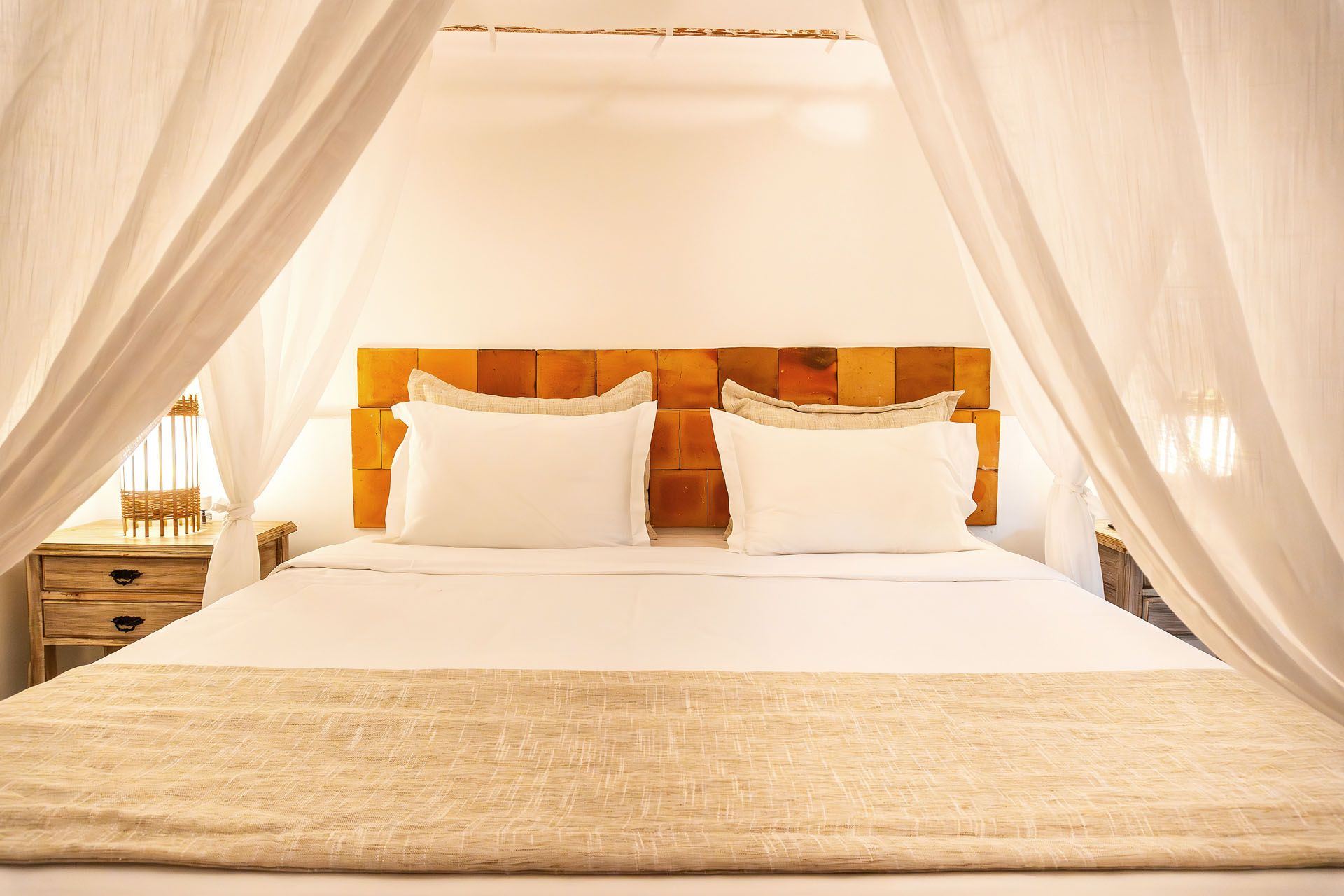 Cama com dossel transparente, lençóis brancos e cobertor bege em um quarto iluminado.
