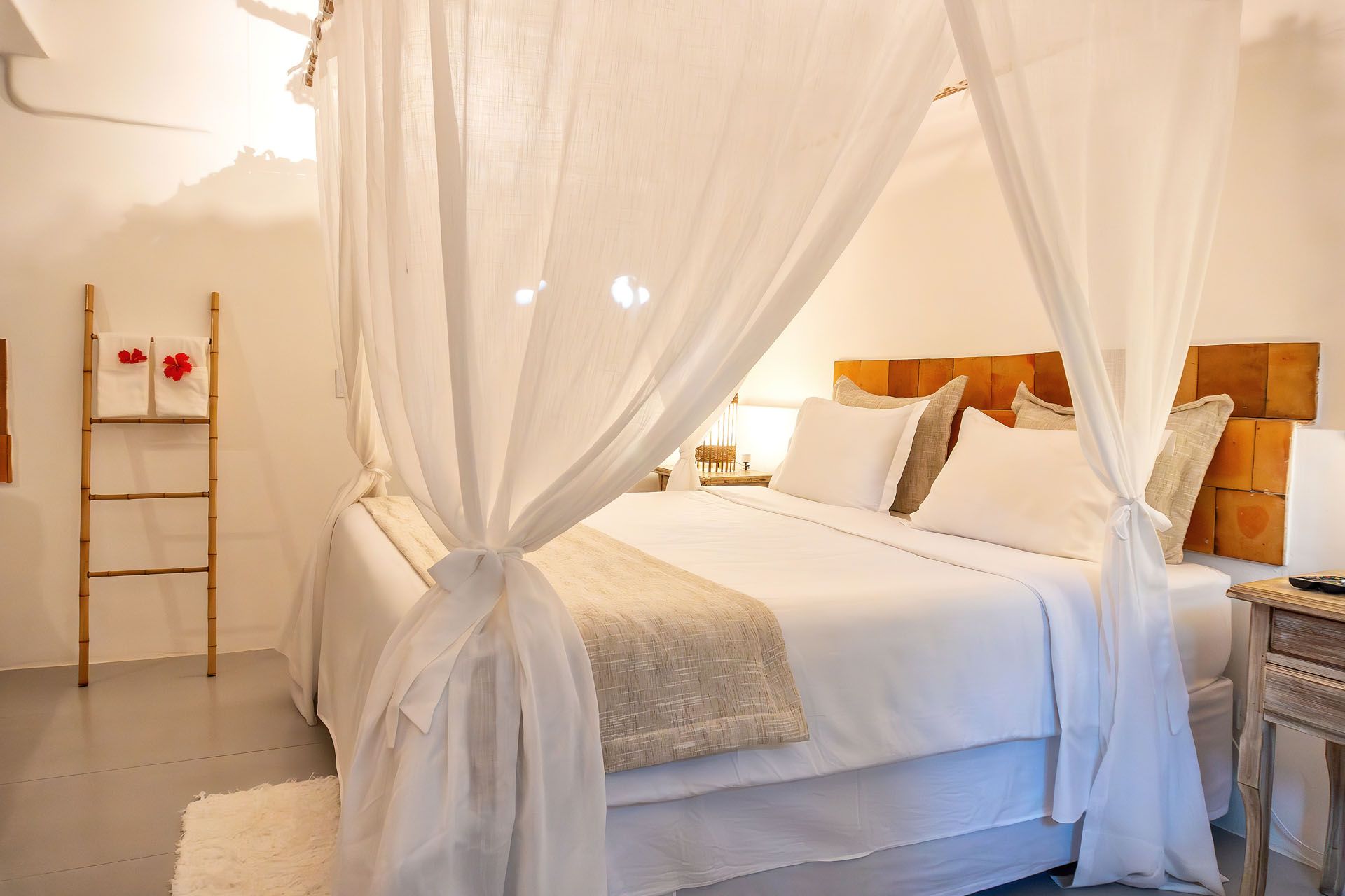 Quarto aconchegante em tons de branco com cortinas de dossel transparentes, cabeceira de madeira e abajures com luz quente.