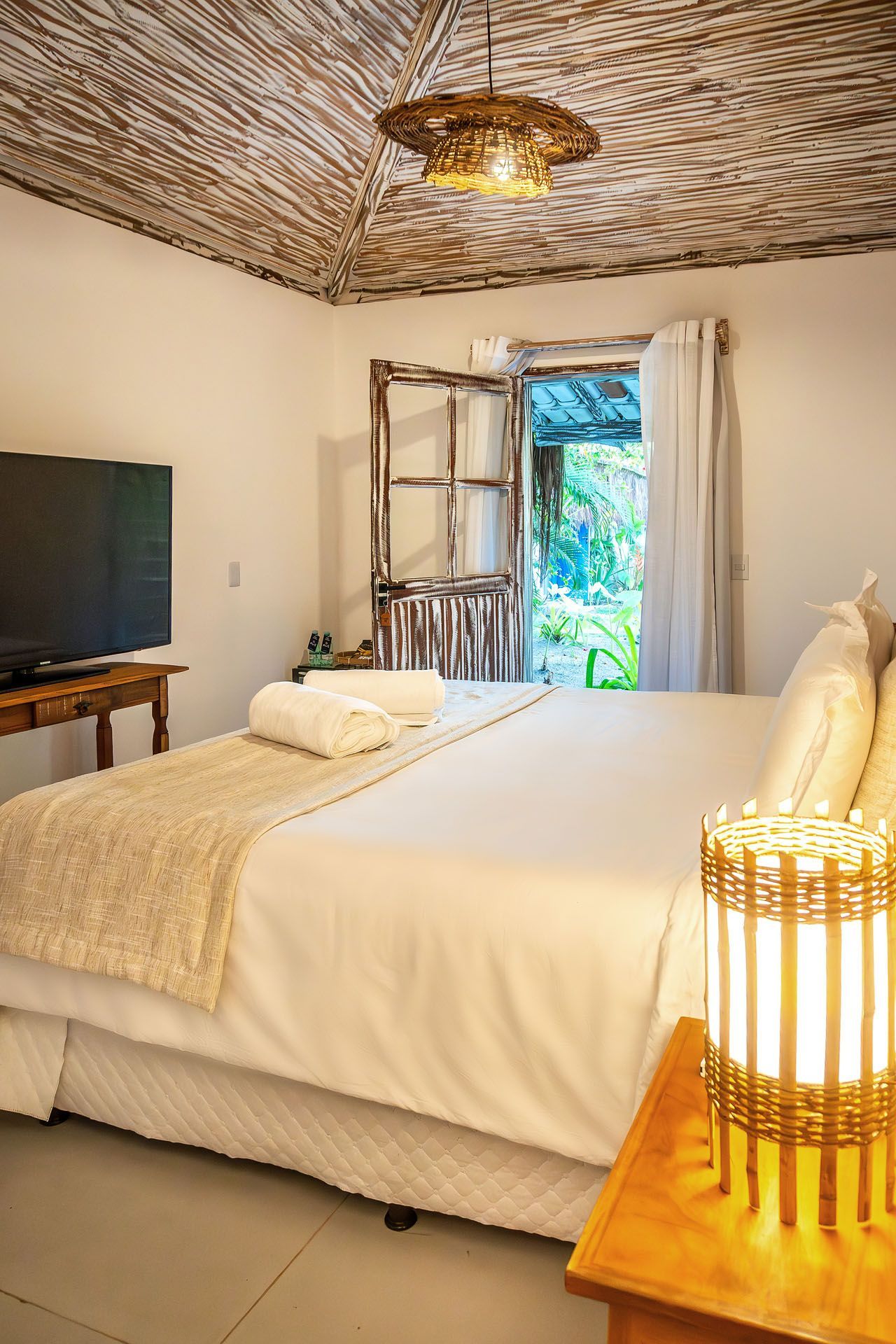Quarto luminoso com cama, luminária de teto de vime e portas de varanda abertas para a vegetação tropical.