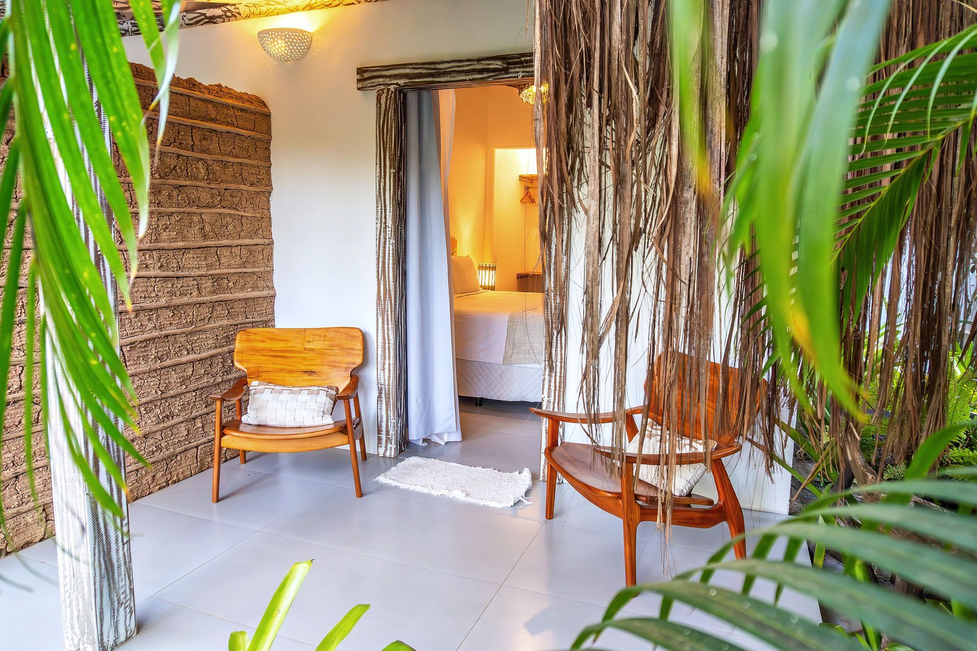 Área de estar tropical no pátio com cadeiras de vime, plantas suspensas e uma porta aberta para um ambiente iluminado.