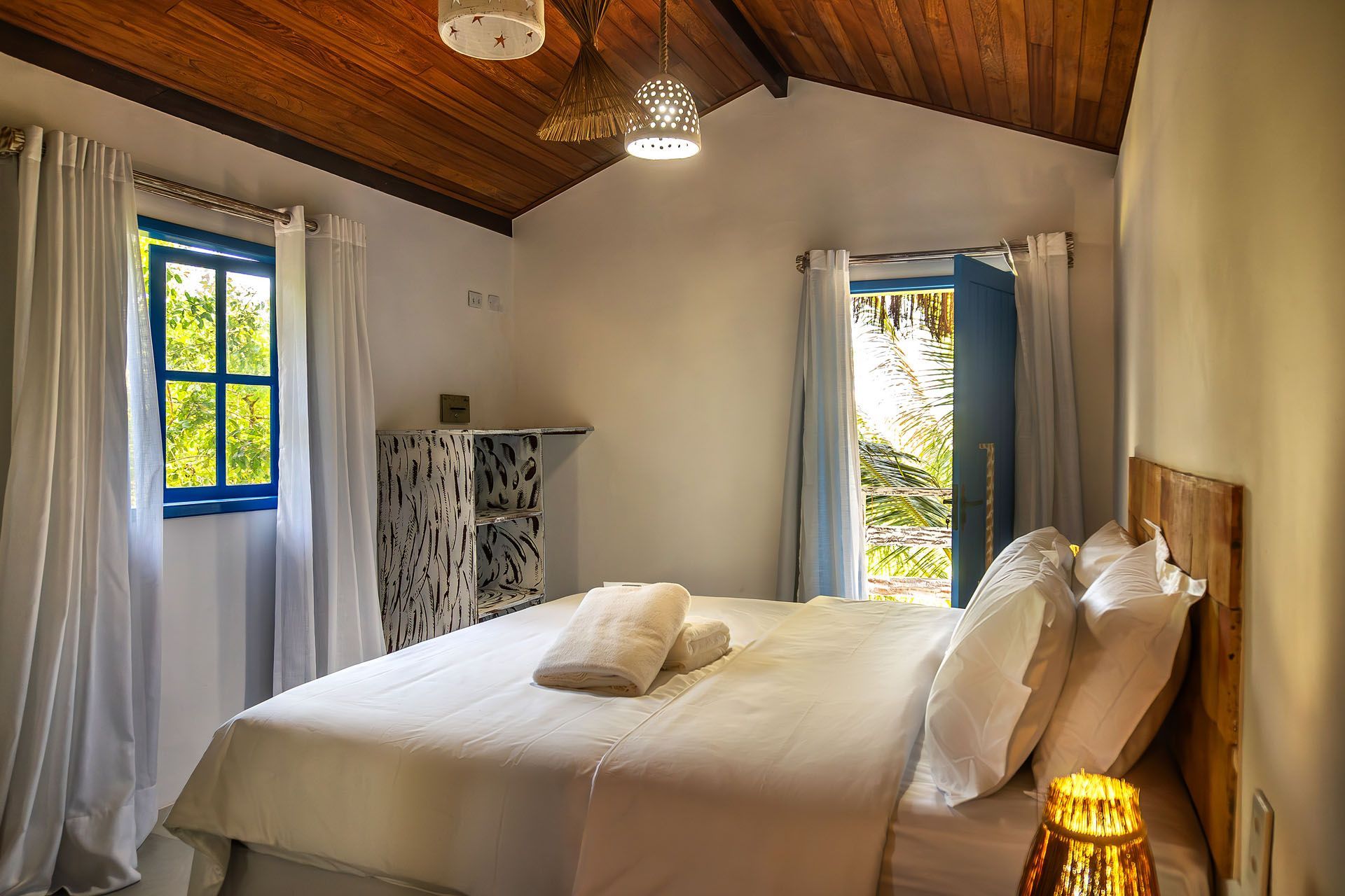 Quarto aconchegante com teto de madeira, cama branca, cortinas azuis e porta aberta para a luz do sol.