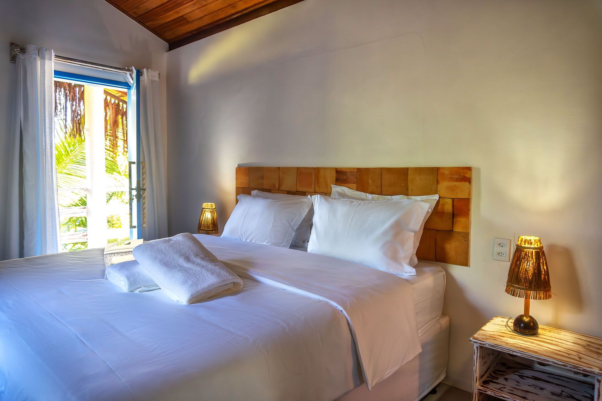 Quarto luminoso com roupa de cama branca, cabeceira de madeira, candeeiros de cabeceira e janela com cortinas.