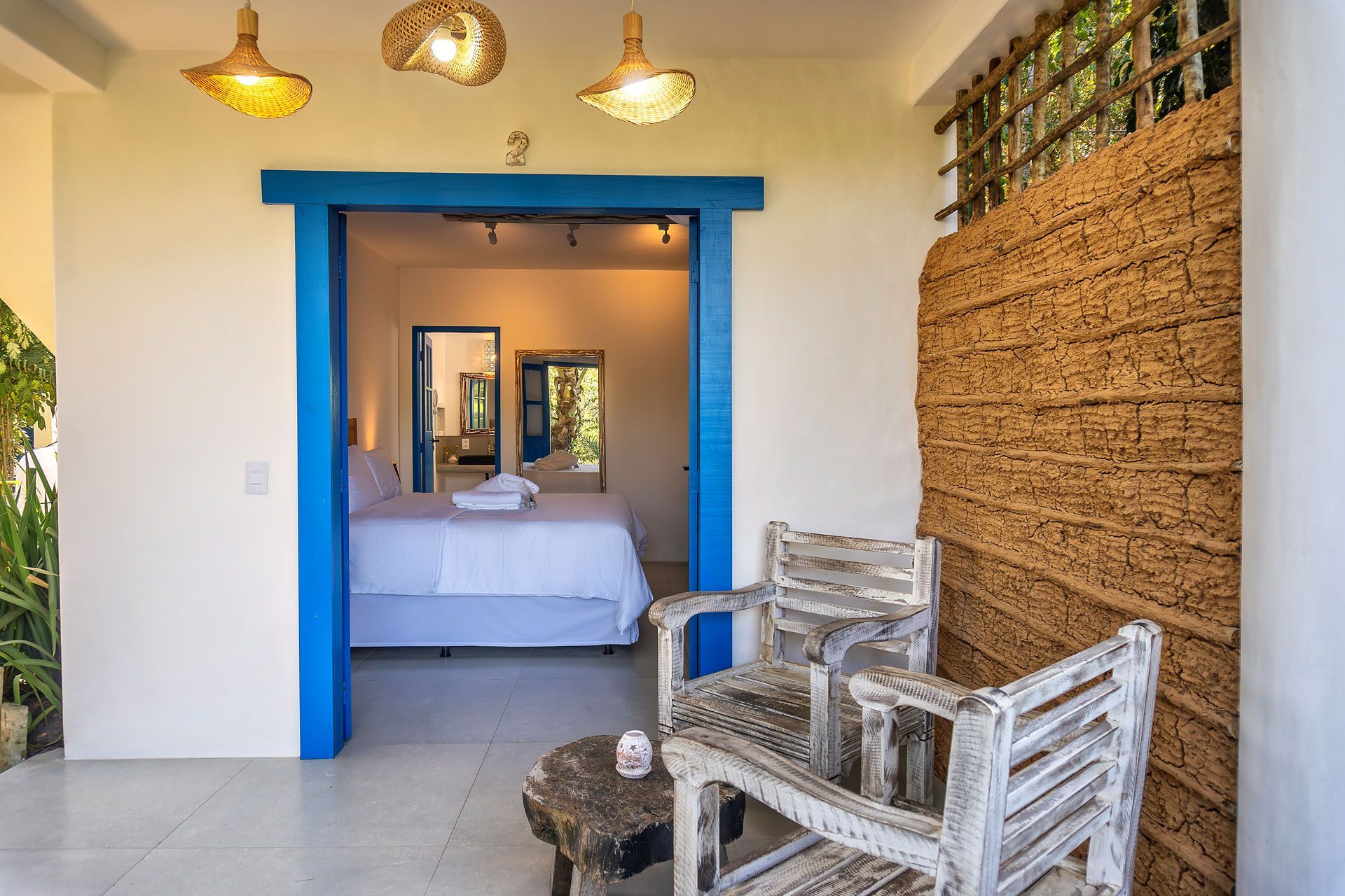 Área de estar com assentos ao lado de uma porta com moldura azul que dá acesso a um quarto, com decoração de parede em tecido e luminárias pendentes.