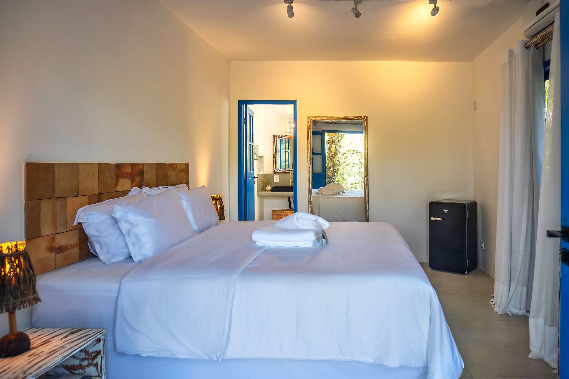 Quarto luminoso com uma cama grande, lençóis brancos, espelho com moldura azul e porta aberta para um pátio ensolarado.