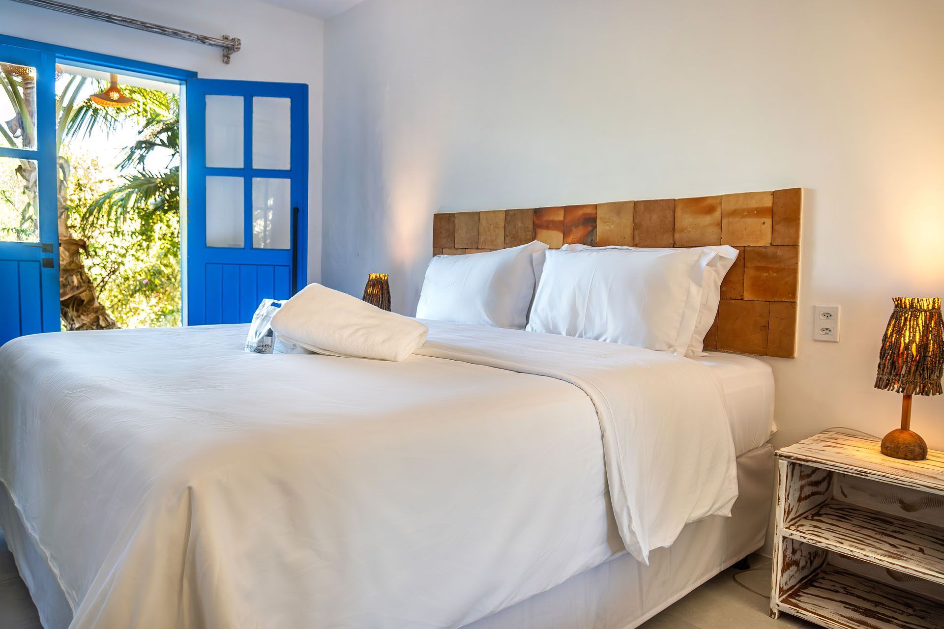 Quarto luminoso com cama branca, persianas azuis e janela aberta com vista para a vegetação tropical.