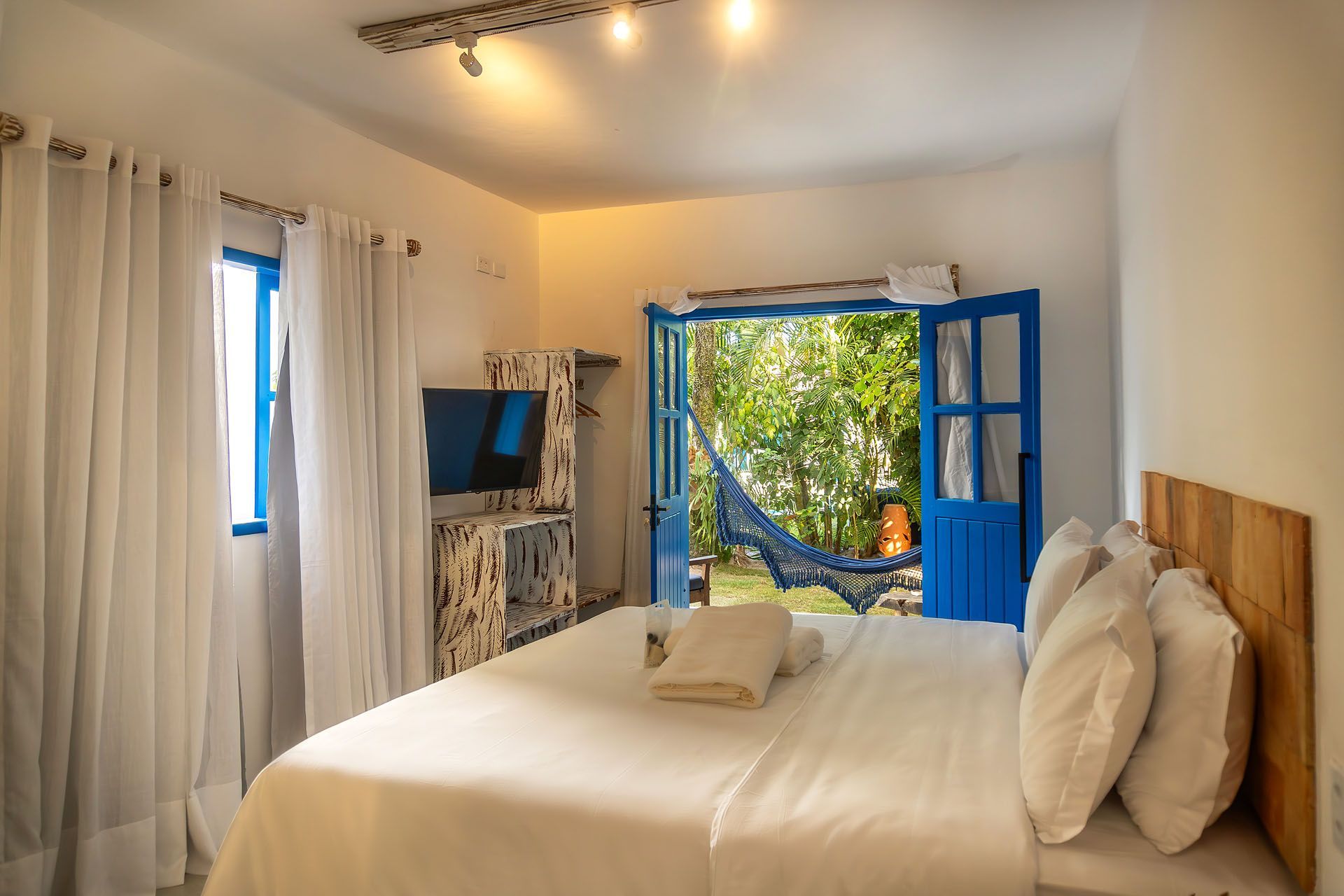 Quarto luminoso com cama branca, portas azuis, janela aberta e muita luz solar.