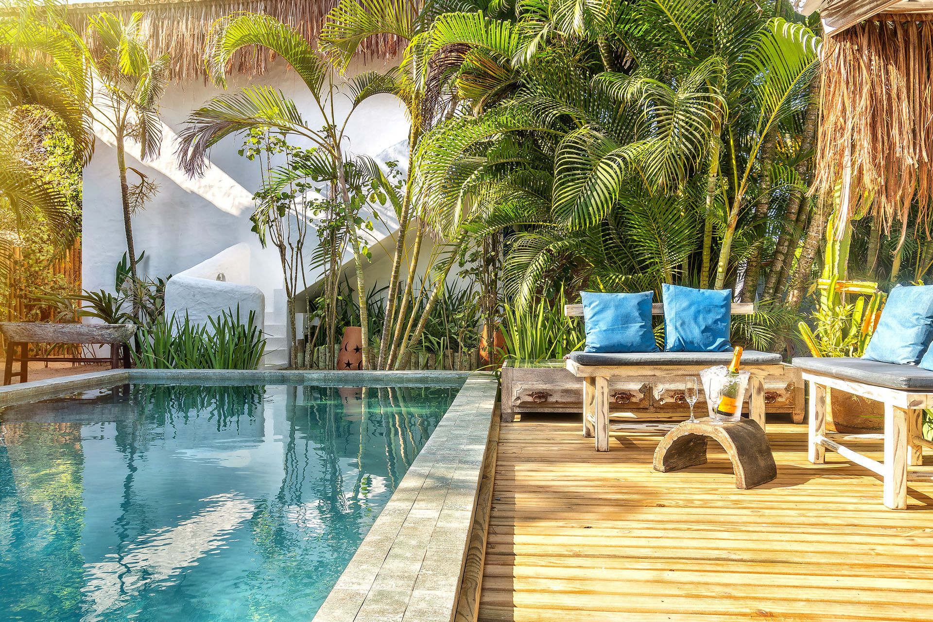 Deck tropical à beira da piscina com almofadas azuis, palmeiras e uma piscina turquesa.