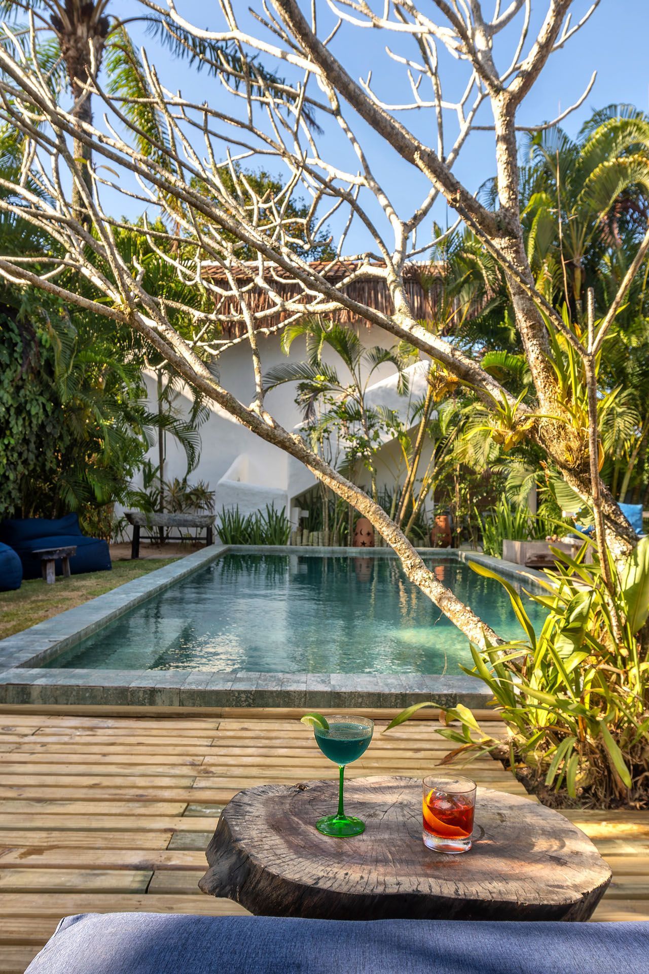 Dois coquetéis em uma mesa de pátio ao lado da piscina, com árvores e uma casa branca ao fundo.