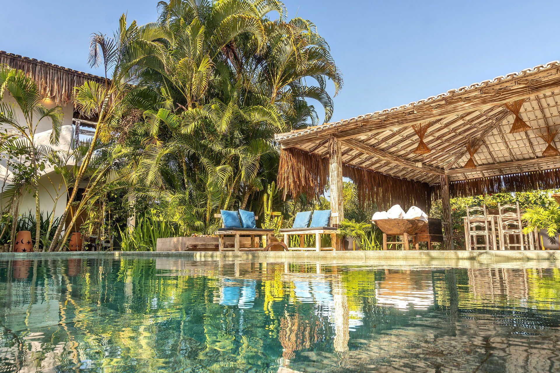 Piscina de resort tropical com palmeiras, cabana de palha e reflexos do sol na água cristalina.