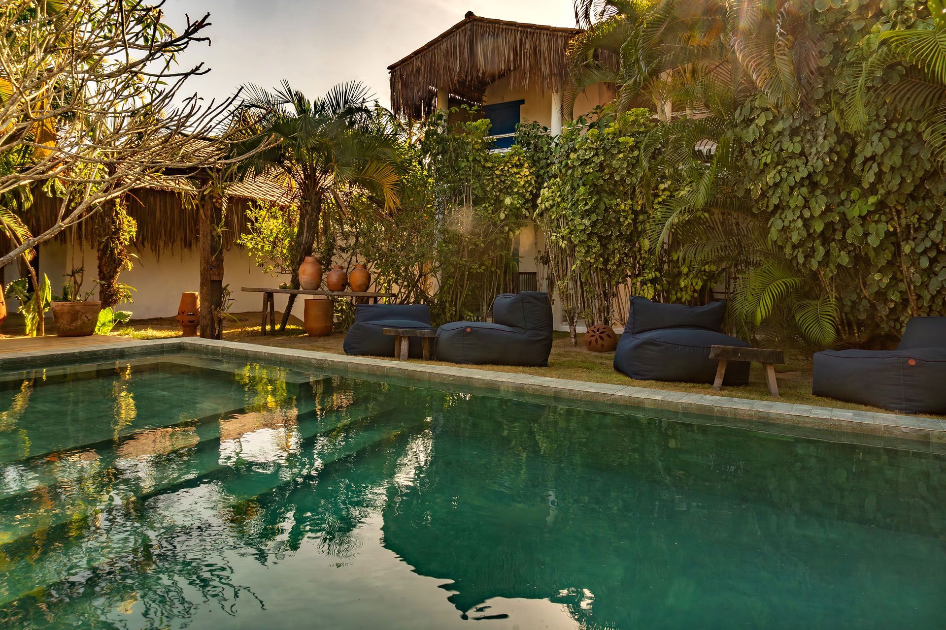 Piscina tropical no pátio, com vegetação exuberante, espreguiçadeiras e luz solar quente refletida.