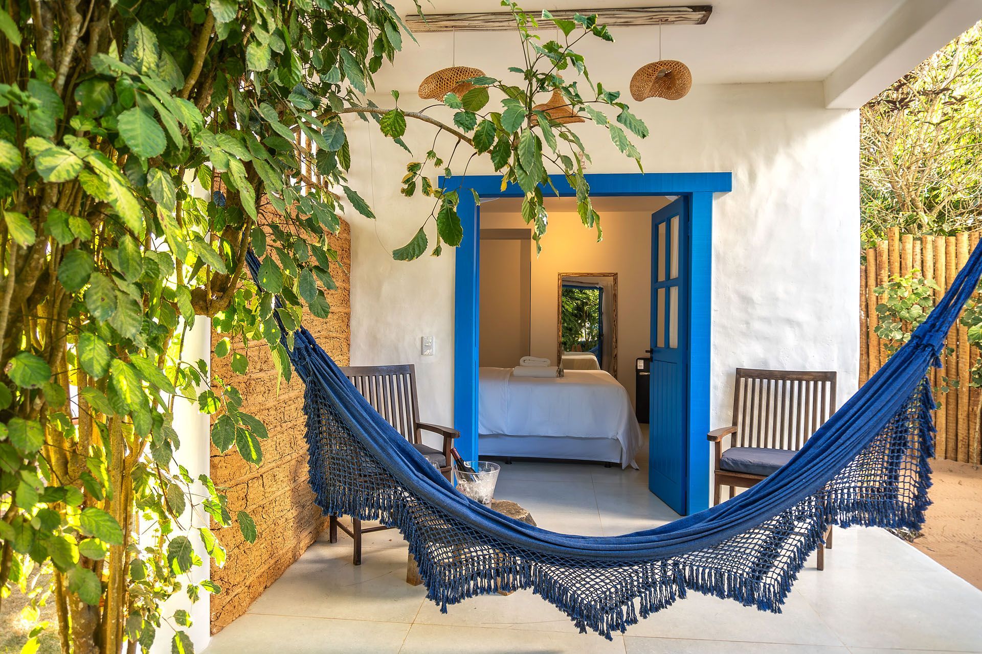 Rede azul em um pátio ensolarado com trepadeiras frondosas e uma porta que emoldura uma cama interna.