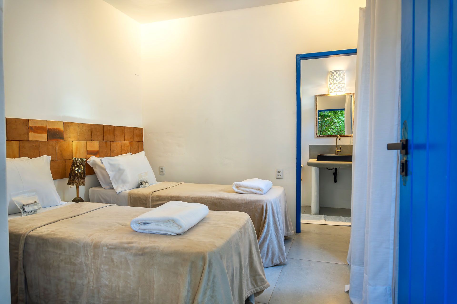 Quarto pequeno com duas camas de solteiro, lençóis bege e uma porta azul que dá acesso ao banheiro.