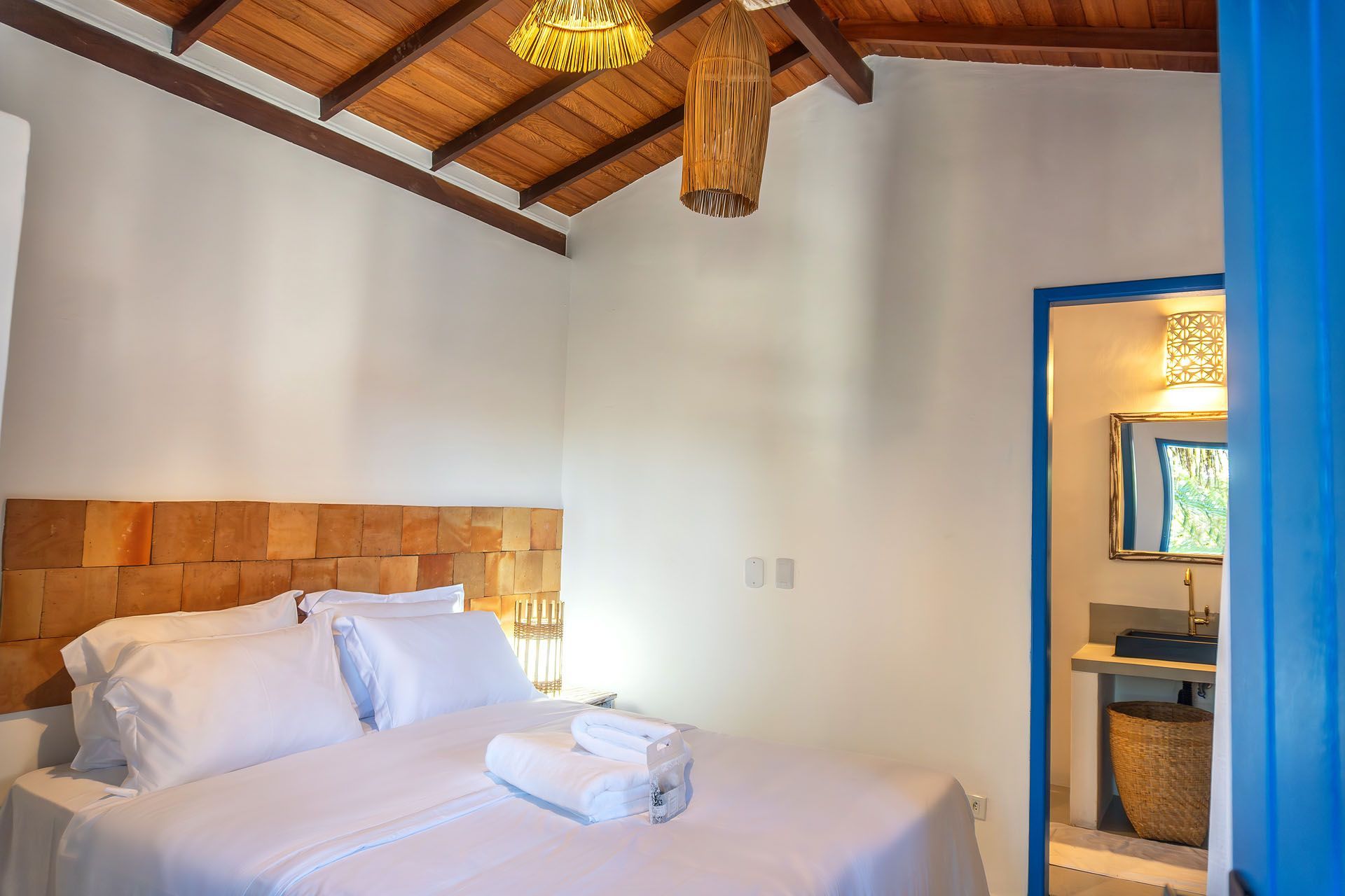 Quarto luminoso com cama branca, cabeceira de madeira e vigas expostas no teto, com acesso a um banheiro com azulejos azuis.