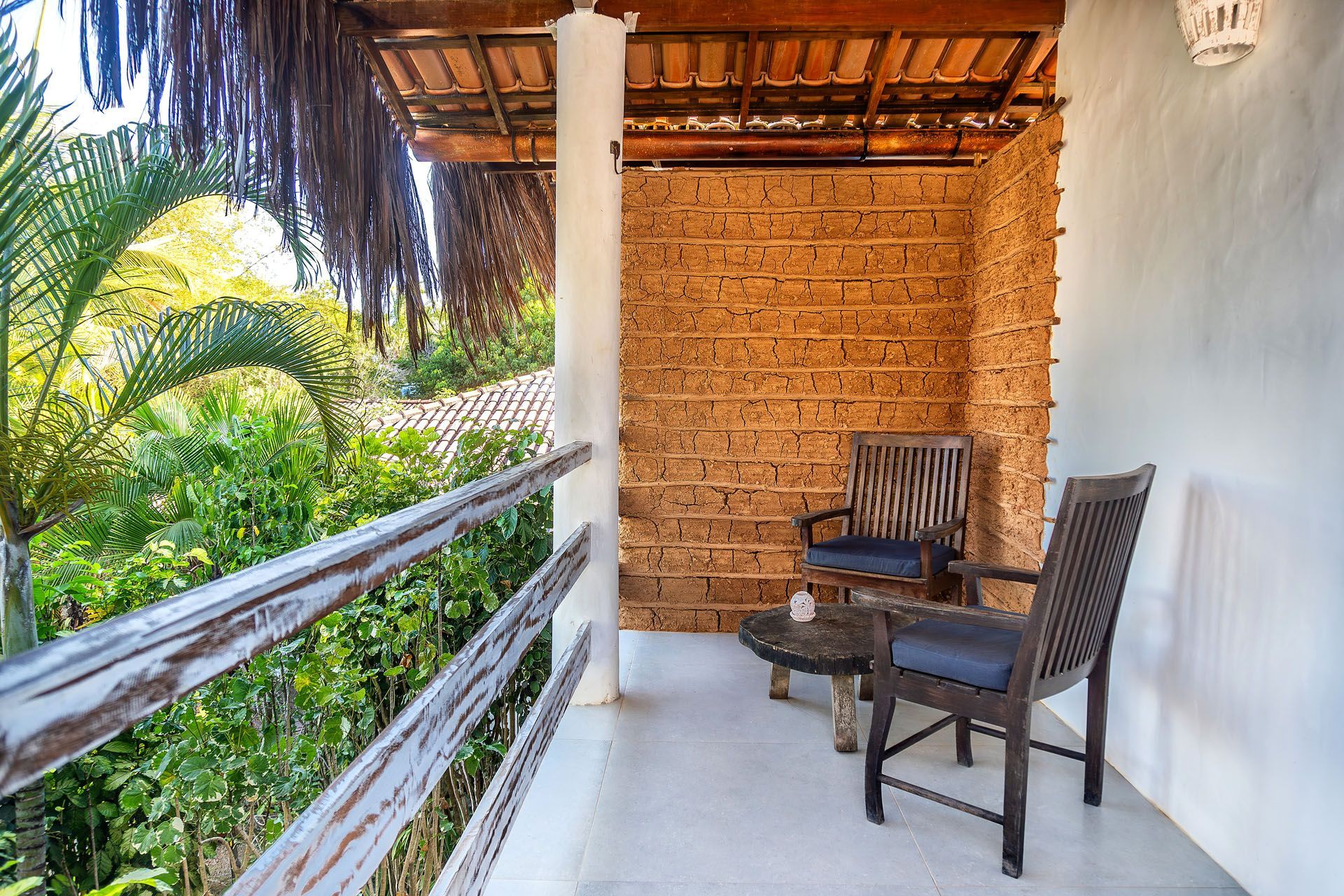 Varanda com duas cadeiras de madeira, uma pequena mesa e vegetação tropical além do parapeito.