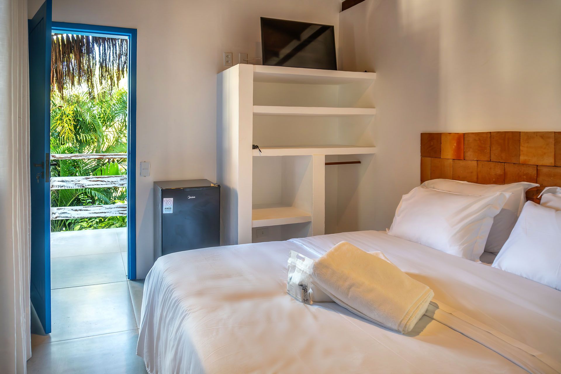 Quarto luminoso com cama branca, prateleiras abertas e porta aberta para uma vista verdejante do exterior.