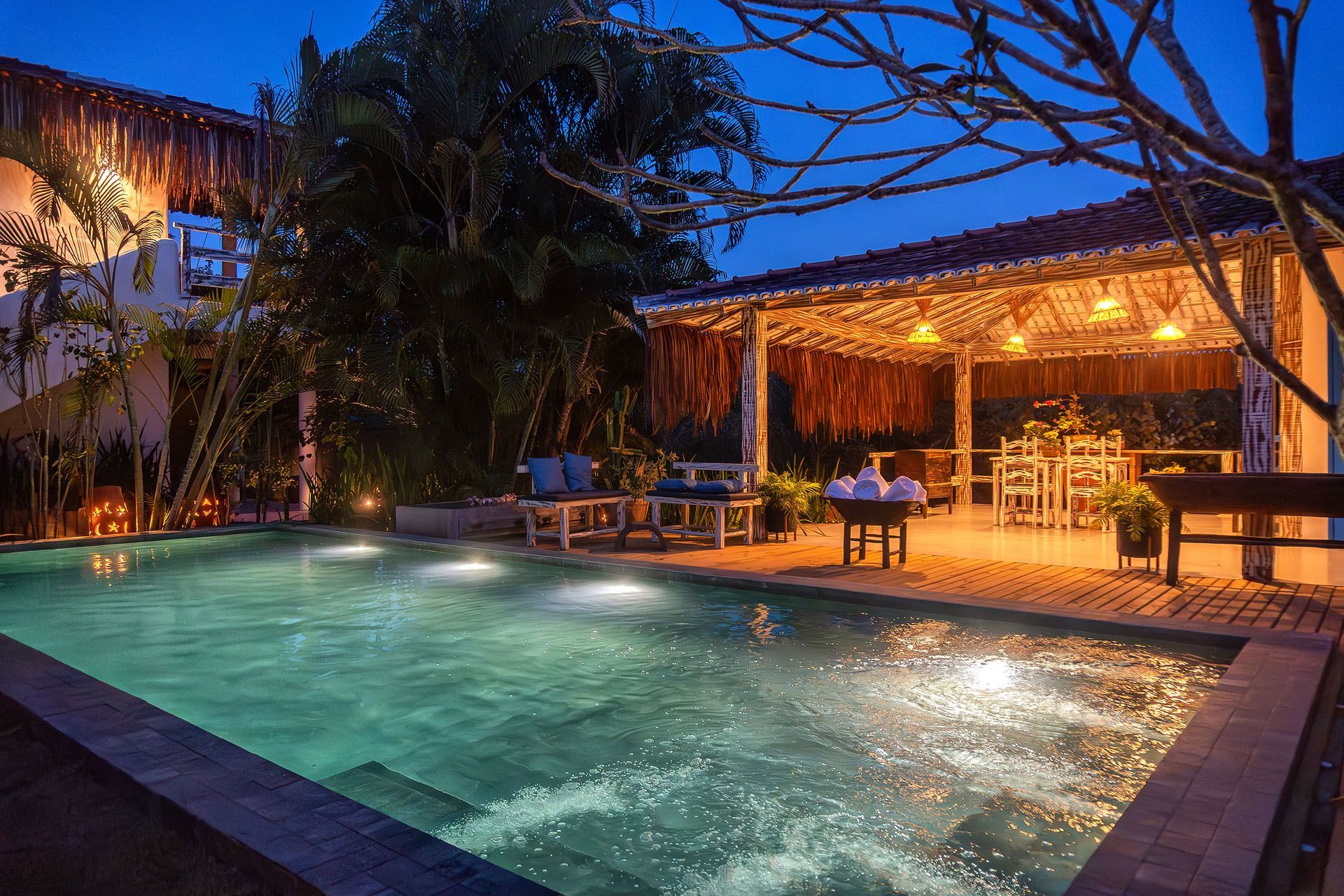 Piscina iluminada do resort ao entardecer, com espreguiçadeiras e luzes aconchegantes sob um telhado de palha.