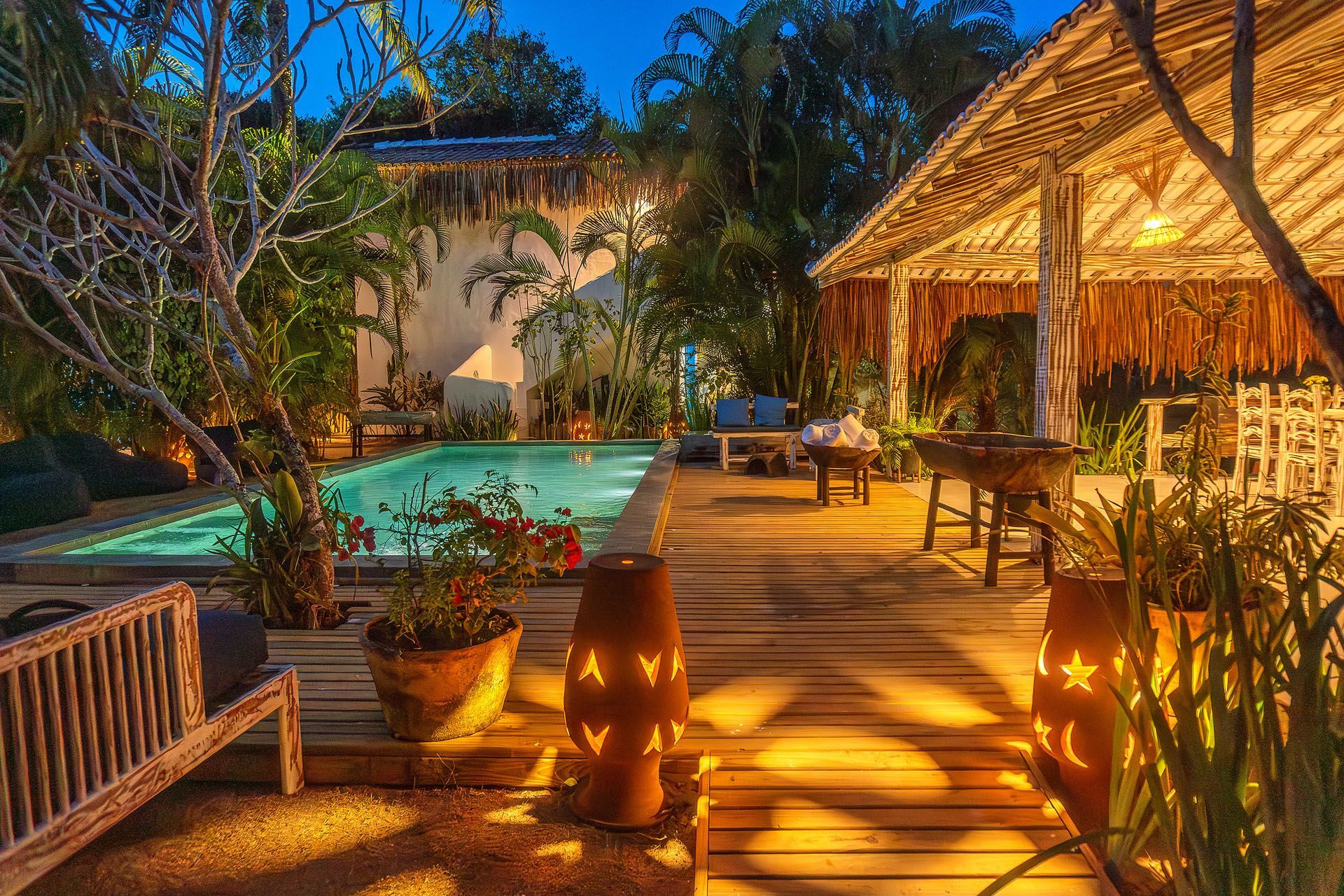 Piscina de resort tropical ao entardecer, com deck de madeira, pavilhão de palha e iluminação aconchegante de lanternas.