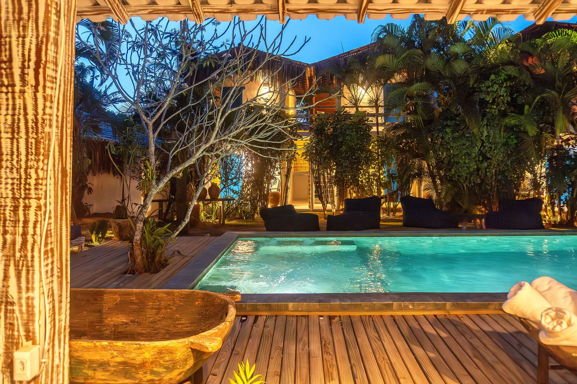 Pátio tropical à beira da piscina ao entardecer, com água turquesa, luzes aconchegantes e vegetação exuberante ao redor de um deck de madeira.